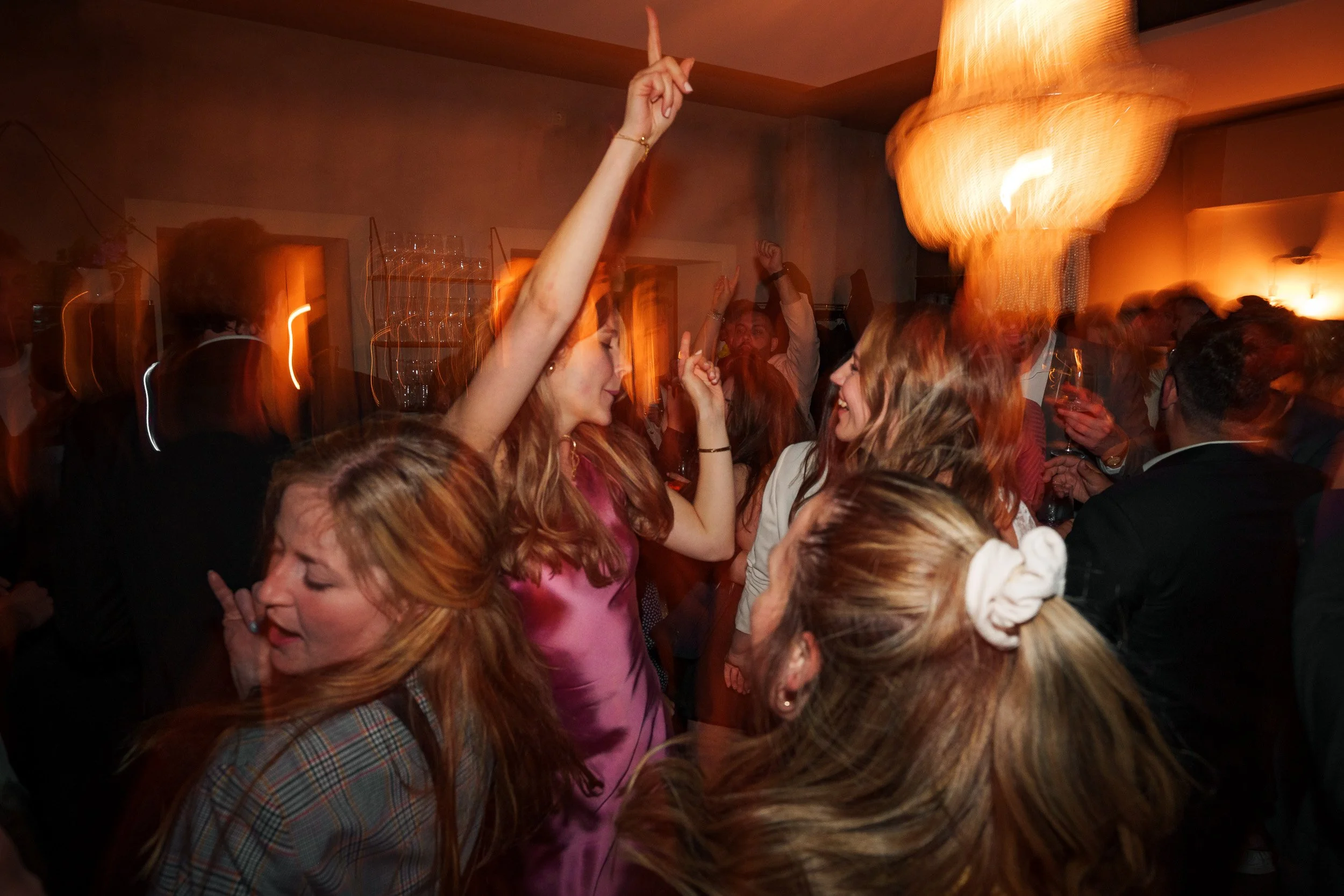Menschen tanzen und feiern auf einer Party, mit Bewegungsunschärfe und warmem Licht, im Hintergrund eine große Lampe.