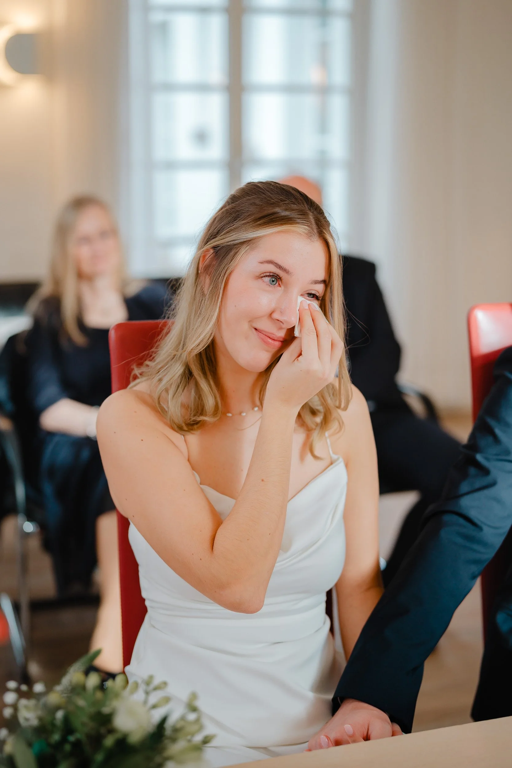 Eine junge Frau in weißem Kleid trauert bei einer Feier, weint und hält sich eine Träne weg. Im Hintergrund sitzen andere Menschen.