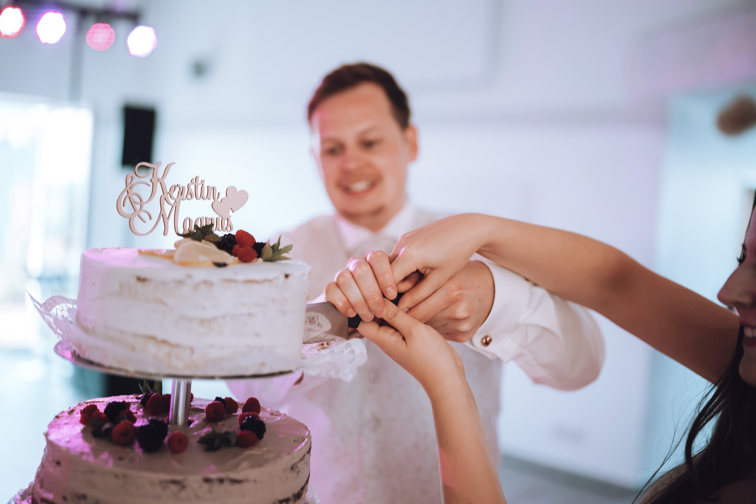 Ein Brautpaar schneidet gemeinsam eine Hochzeitstorte auf einer Feier, die Torte ist geschichtet mit Beeren und hat einen Namen auf einem Dekorationsschild.