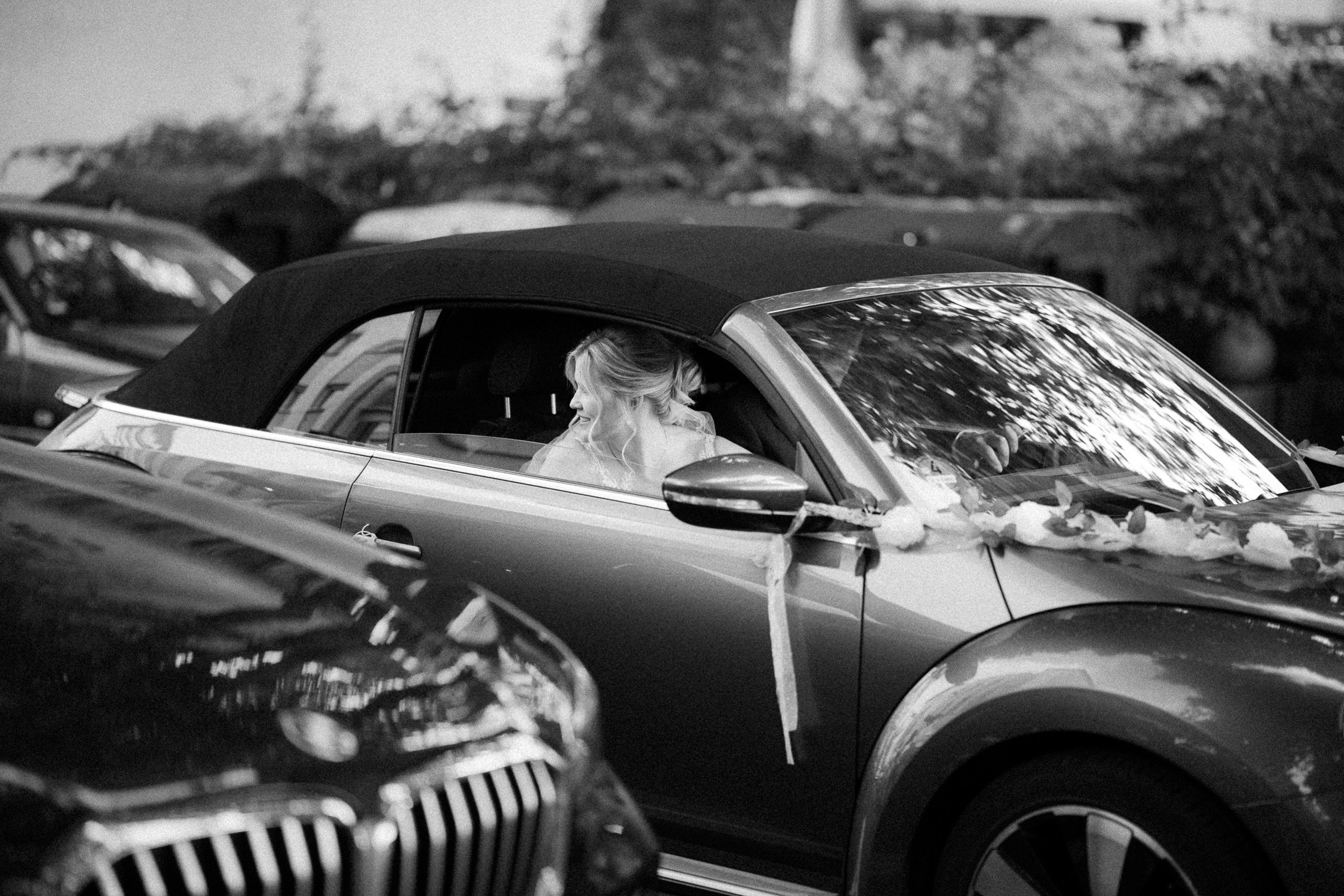 Eine Frau sitzt in einem Cabrio mit schwarz-weißem Verdeck, dekoriert für eine Feier, und schaut nach rechts. Das Auto ist gedeckt mit Dekorationsmaterialien, wahrscheinlich für eine Hochzeit.