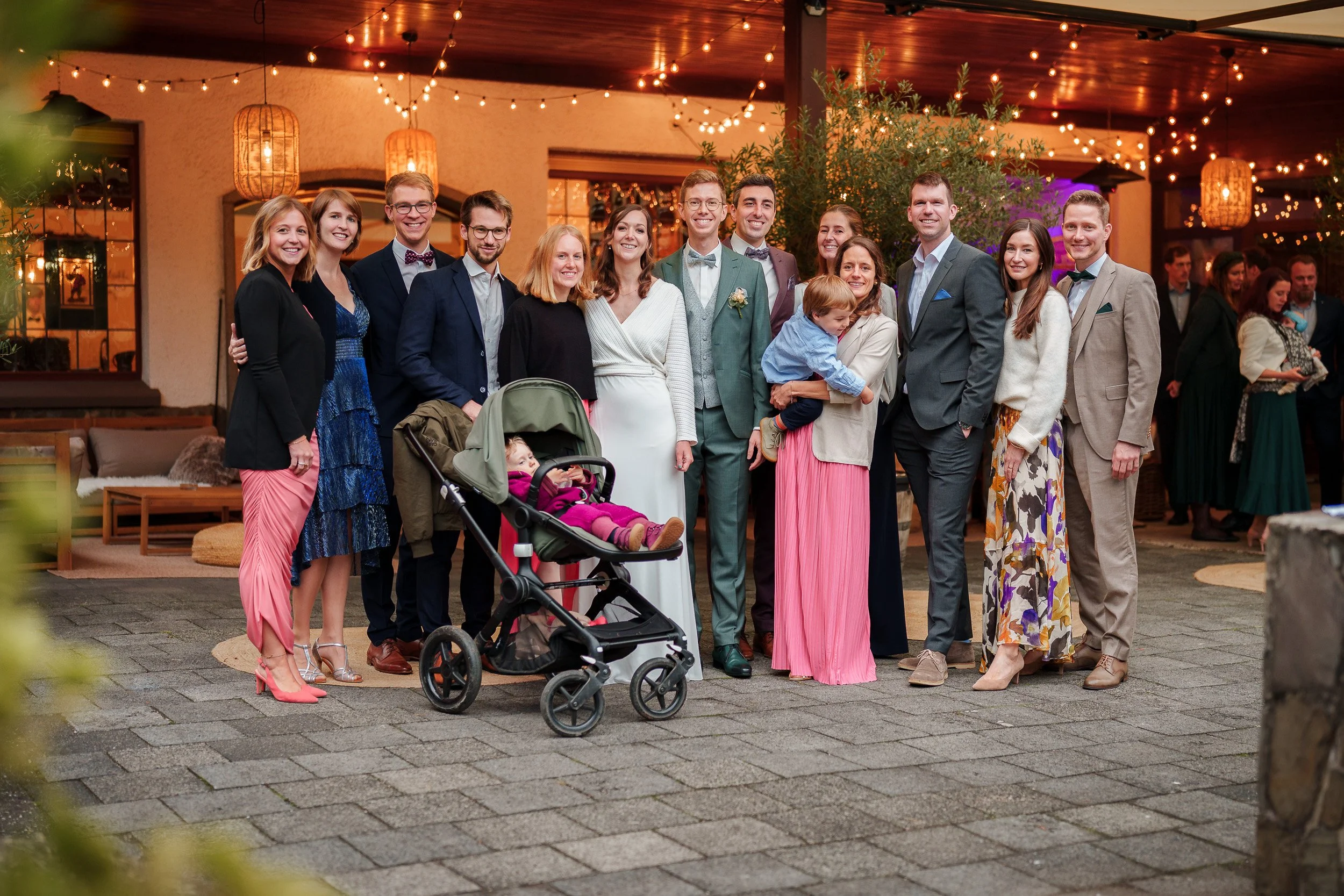 Eine Gruppe von Menschen bei einer Feier, alle lächelnd, im Freien, umgeben von Lichterketten und warmem Licht, mit einem kinderwagen im Vordergrund.