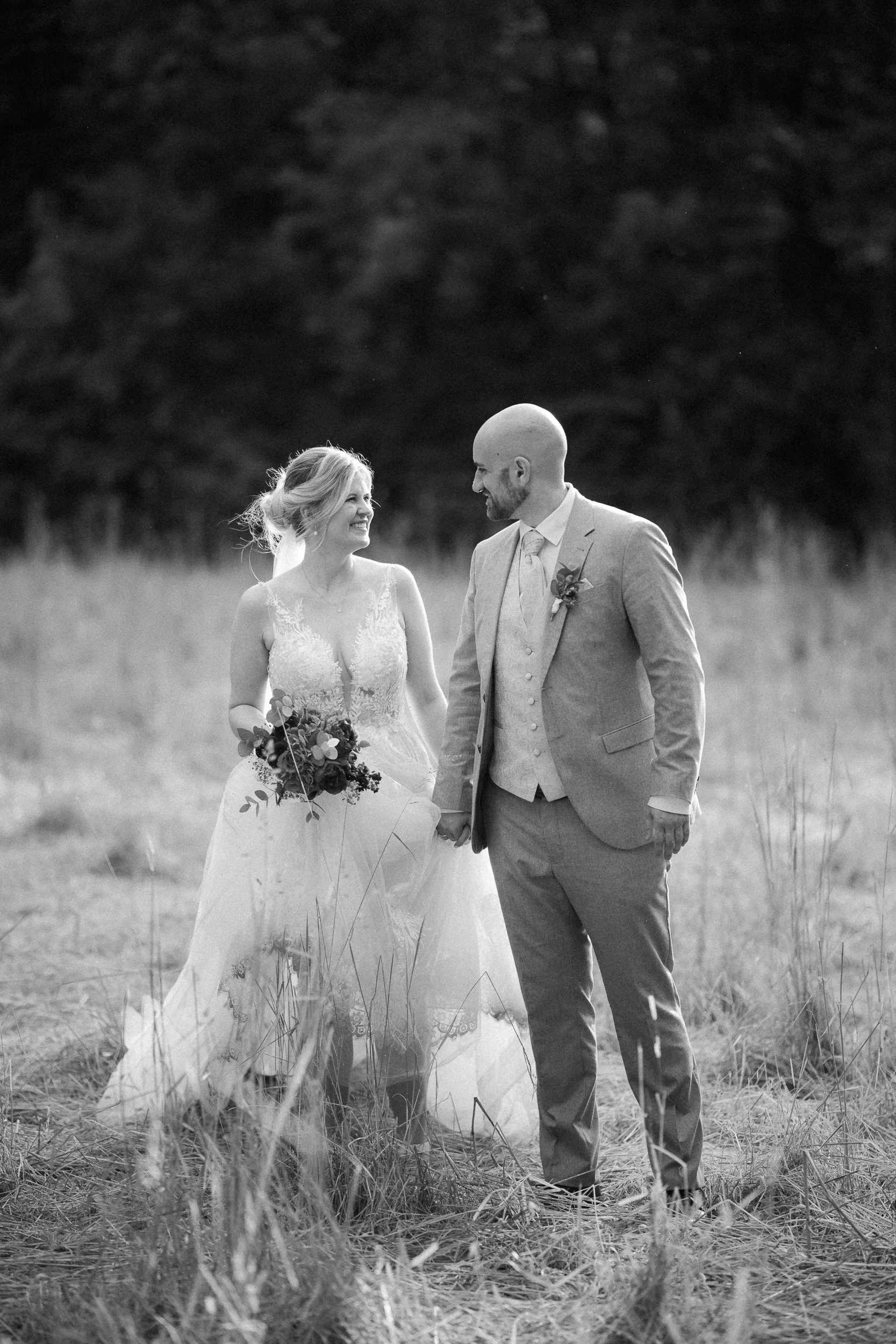 Braut und Bräutigam halten Händchen und schauen sich liebevoll an, auf einer Wiese im Freien bei Sonnenuntergang, schwarz-weiße Hochzeitsfotografie.