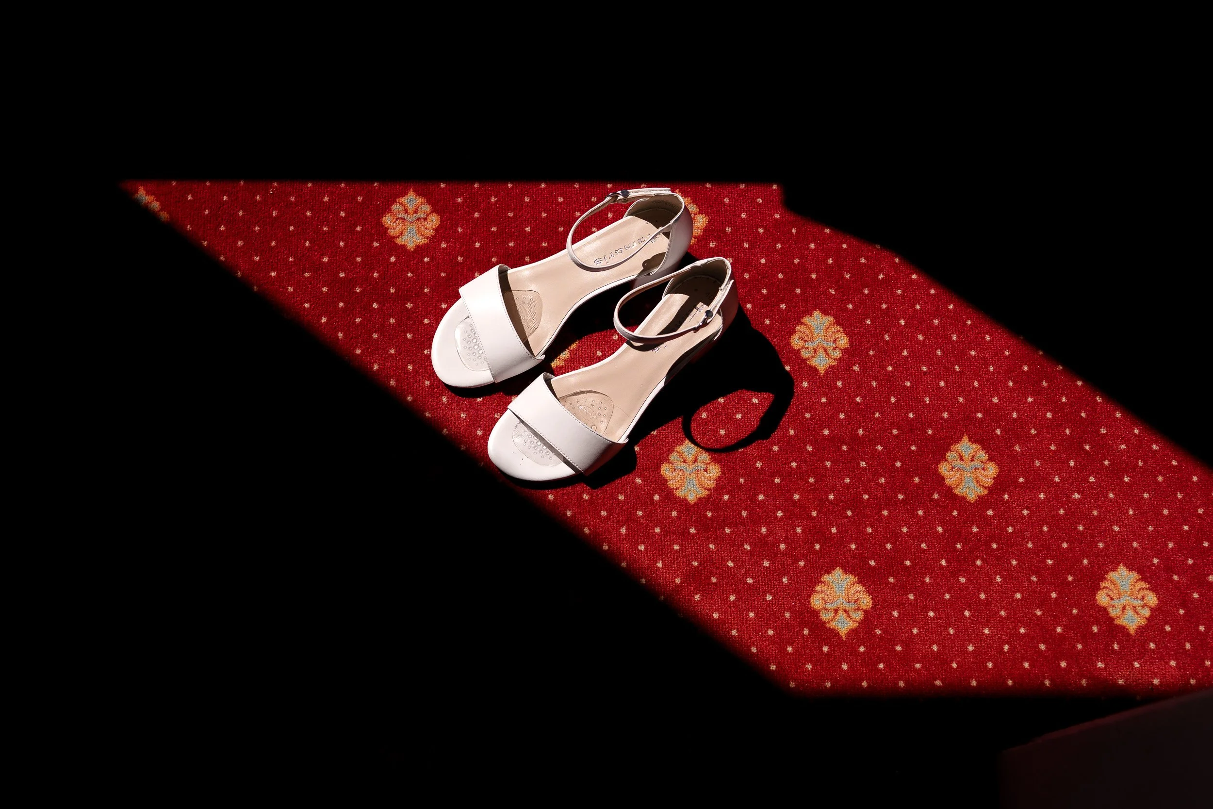 Getting Ready |  Gummersbach | Weiße High Heels auf rotem Teppich mit gold- und blumendekor, beleuchtet von Sonnenlicht und umgeben von Schatten.