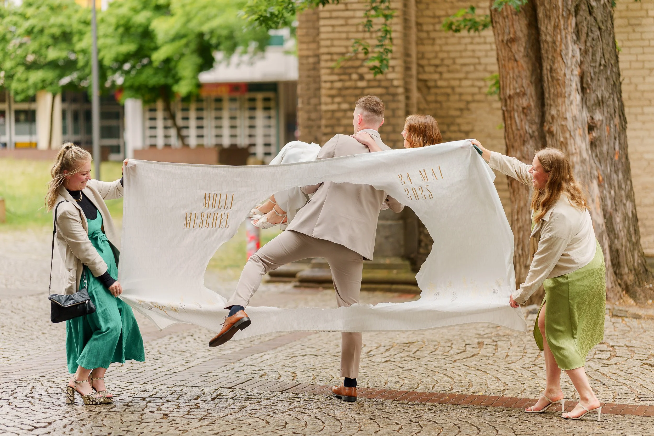 Vier Personen halten ein großes weißes Tuch mit goldener Schrift, auf dem ein Paar liegt, das eine Hochzeitsfeier hat. Zwei Frauen, ein Mann und eine Person in einem Hochzeitsanzug sind draußen auf einem gepflasterten Platz neben Bäumen, die auf eine