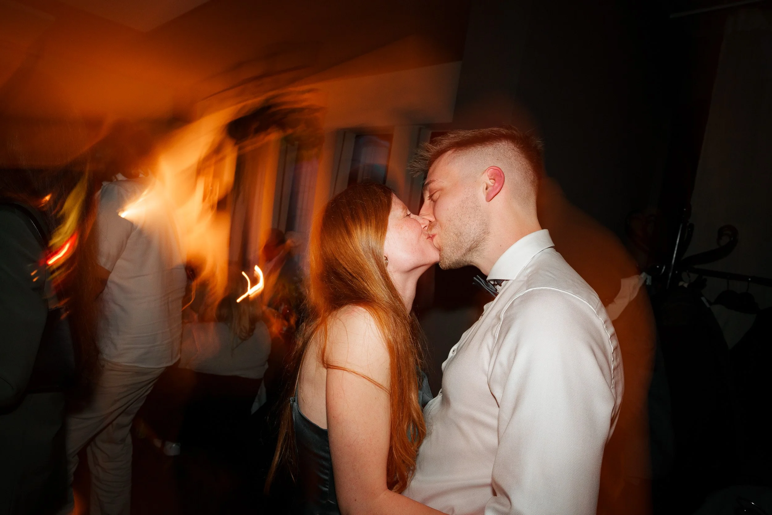 Ein Paar küsst sich bei einer Party in einem dunklen Raum mit warmen Lichtern, im Hintergrund sind andere Gäste und Bewegungsunschärfe