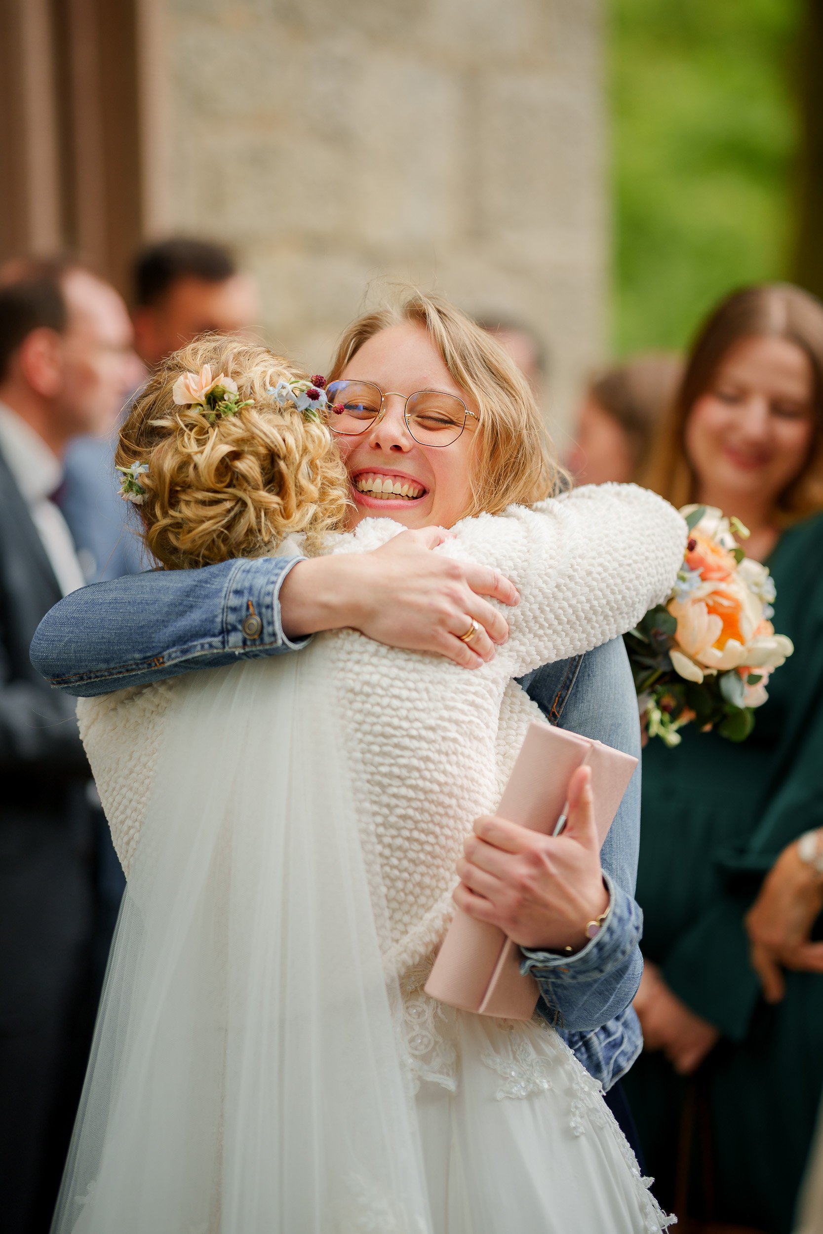 Zwei Frauen umarmen sich herzlich bei einer Hochzeitsfeier. Die lachende Frau trägt Brille und einen weißen Hochzeitskleid, die andere hat lockiges Haar, Blumen im Haar und hält ein kleines Geschenk in der Hand. Im Hintergrund sind weitere Gäste sich