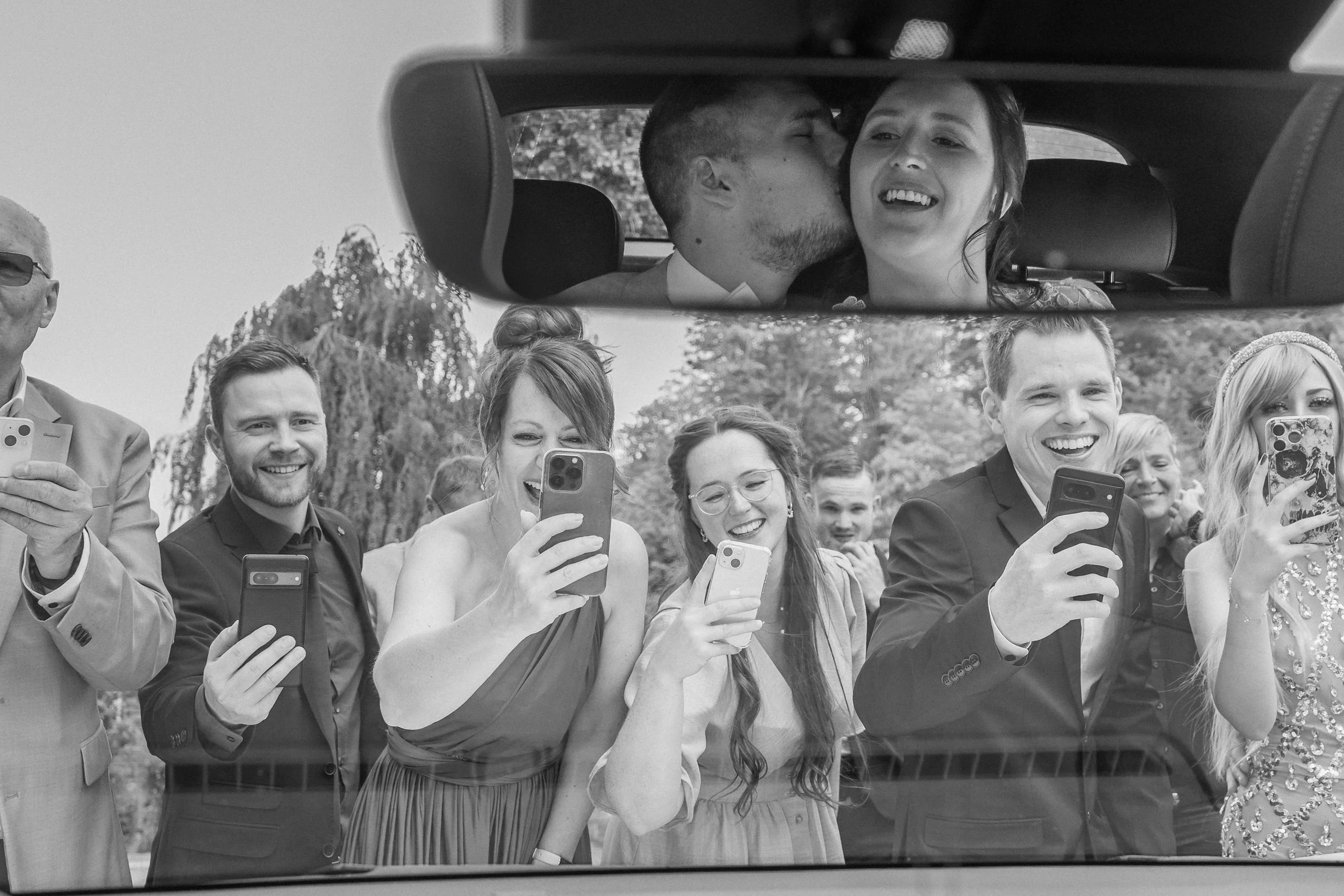 Ein Brautpaar im Hochzeitsauto, umgeben von Freunden, die Fotos mit ihren Handys machen, alle lachen und freuen sich.
