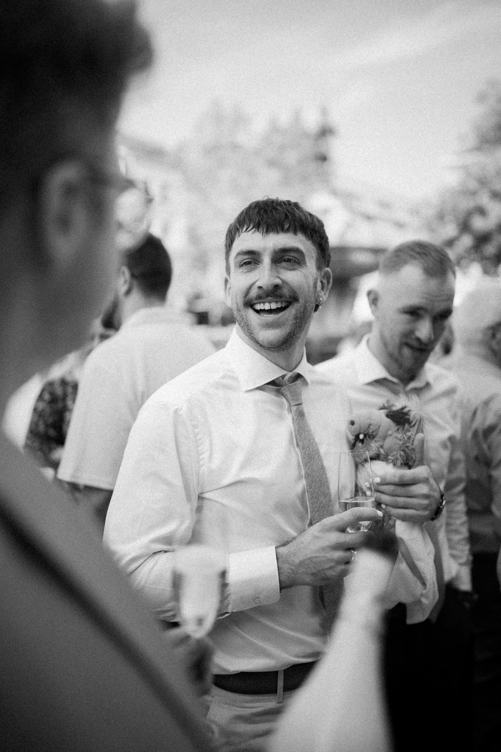 Mann mit dunklem Haar und Bart, lächelnd, hält ein Glas Wein bei einer Feier im Freien, umgeben von anderen Personen.