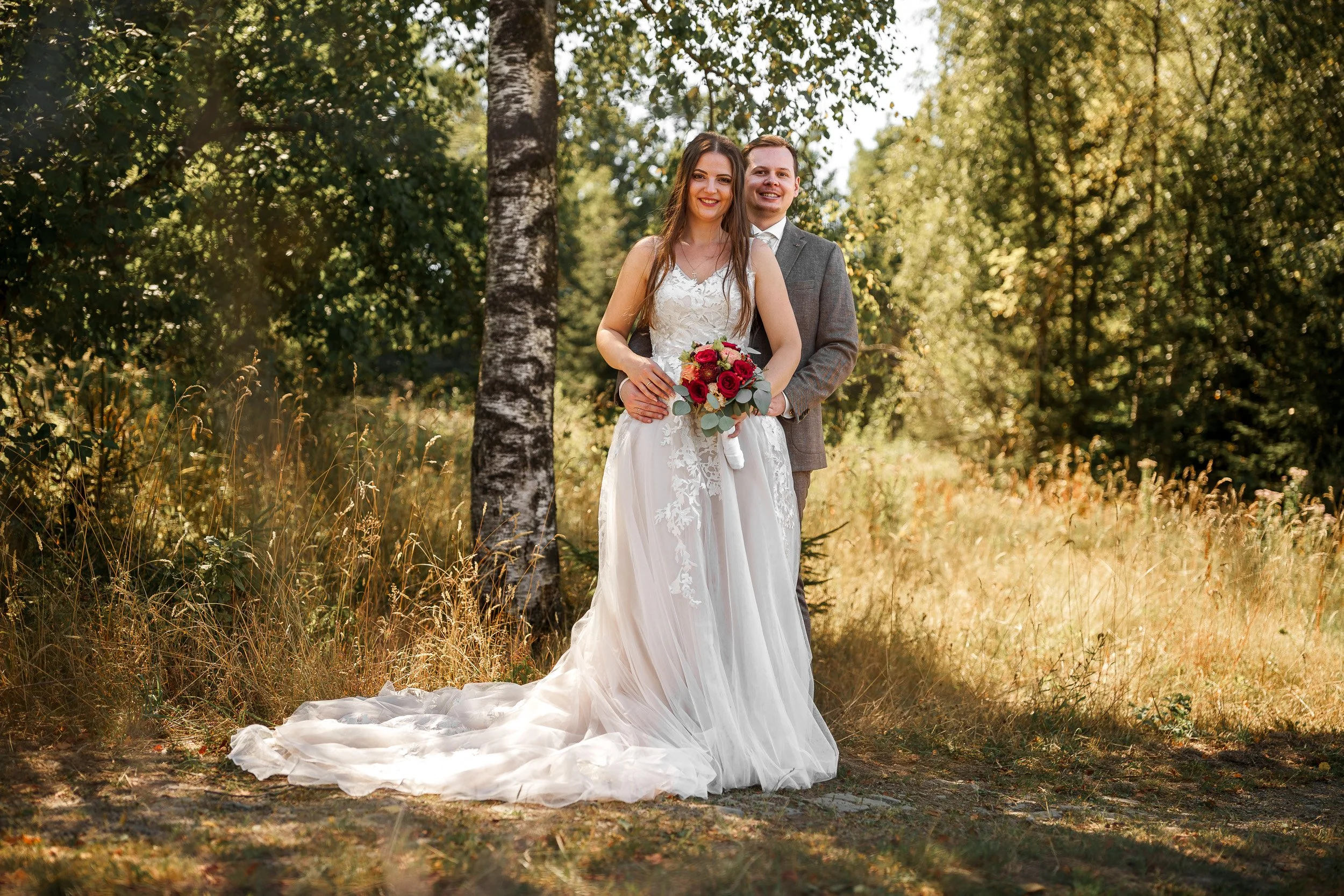 Braut und Bräutigam stehen im Wald, die Braut trägt ein weißes Hochzeitskleid mit Schleier, hält einen Blumenstrauß aus roten und pinken Blumen. Der Bräutigam trägt einen grauen Anzug. Es ist sonnig, das Laub ist grün und die Szenerie wirkt friedlich