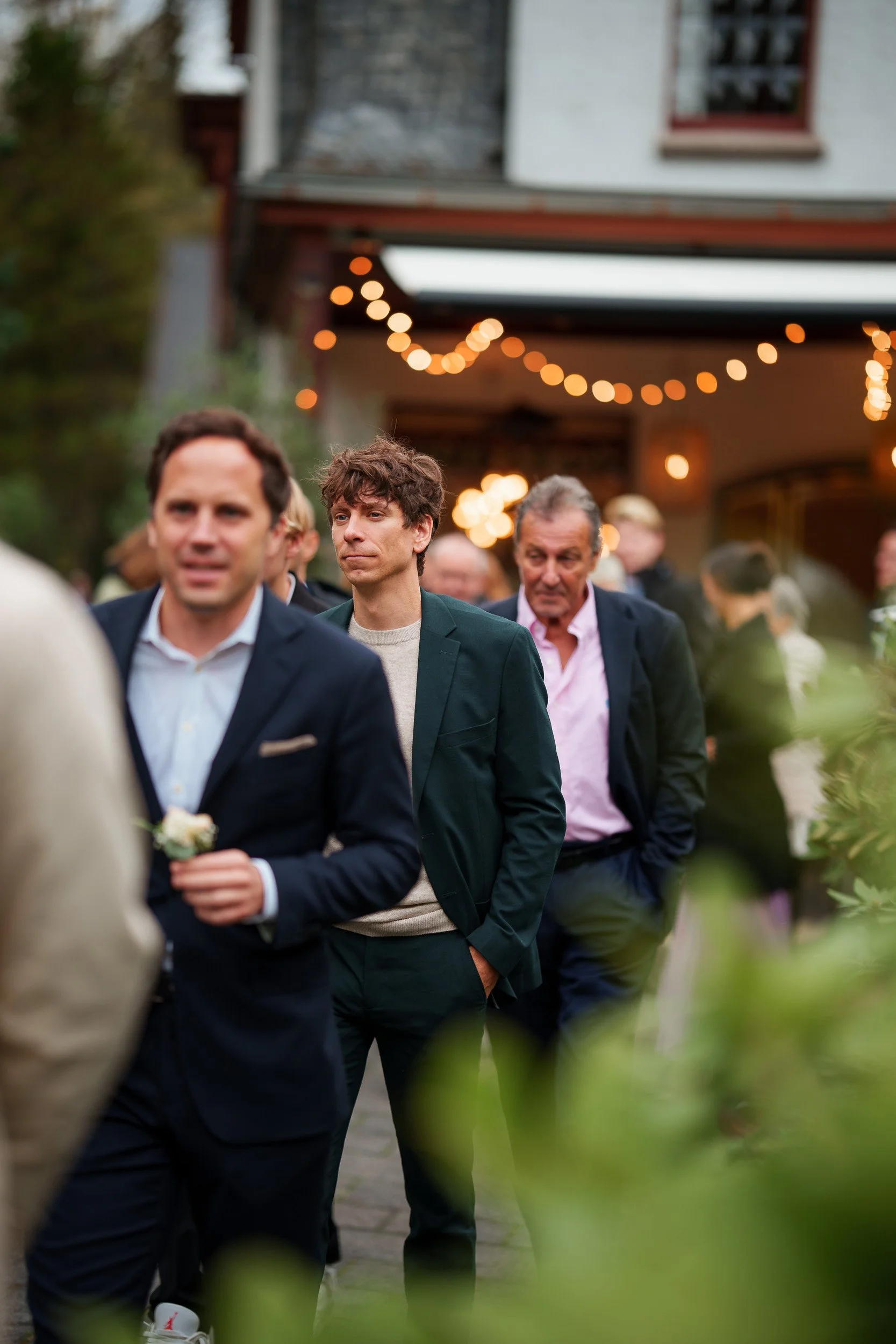 Menschen stehen in einer Warteschlange bei einer Veranstaltung im Freien, im Hintergrund sind Lichterketten und ein Gebäude sichtbar.