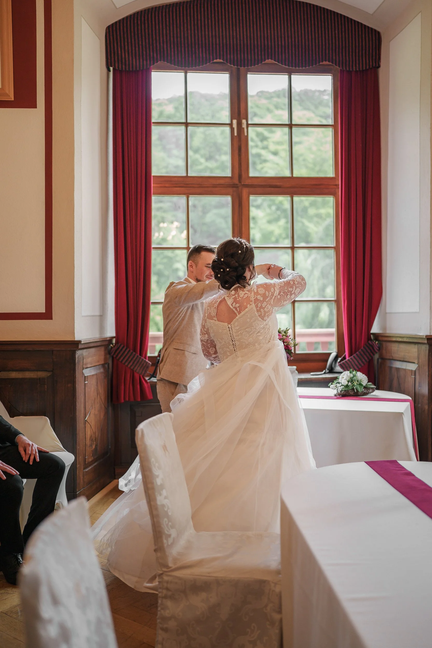 Ein Brautpaar tanzt in einem festlich dekorierten Raum neben einem großen Fenster mit roten Vorhängen.