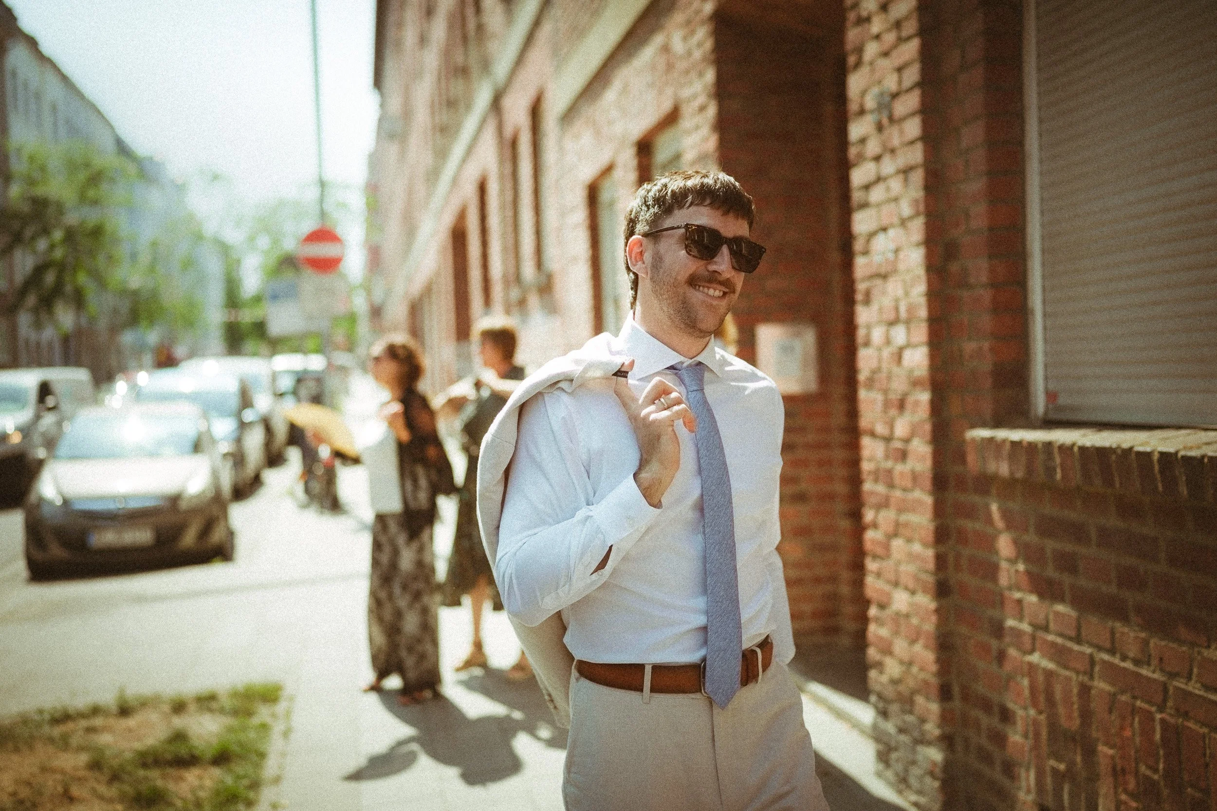Ein lächelnder Mann in einem weißen Hemd und blauer Krawatte, trägt eine Sonnenbrille und eine Jacke über der Schulter, während er auf der Straße in einer Stadt geht.