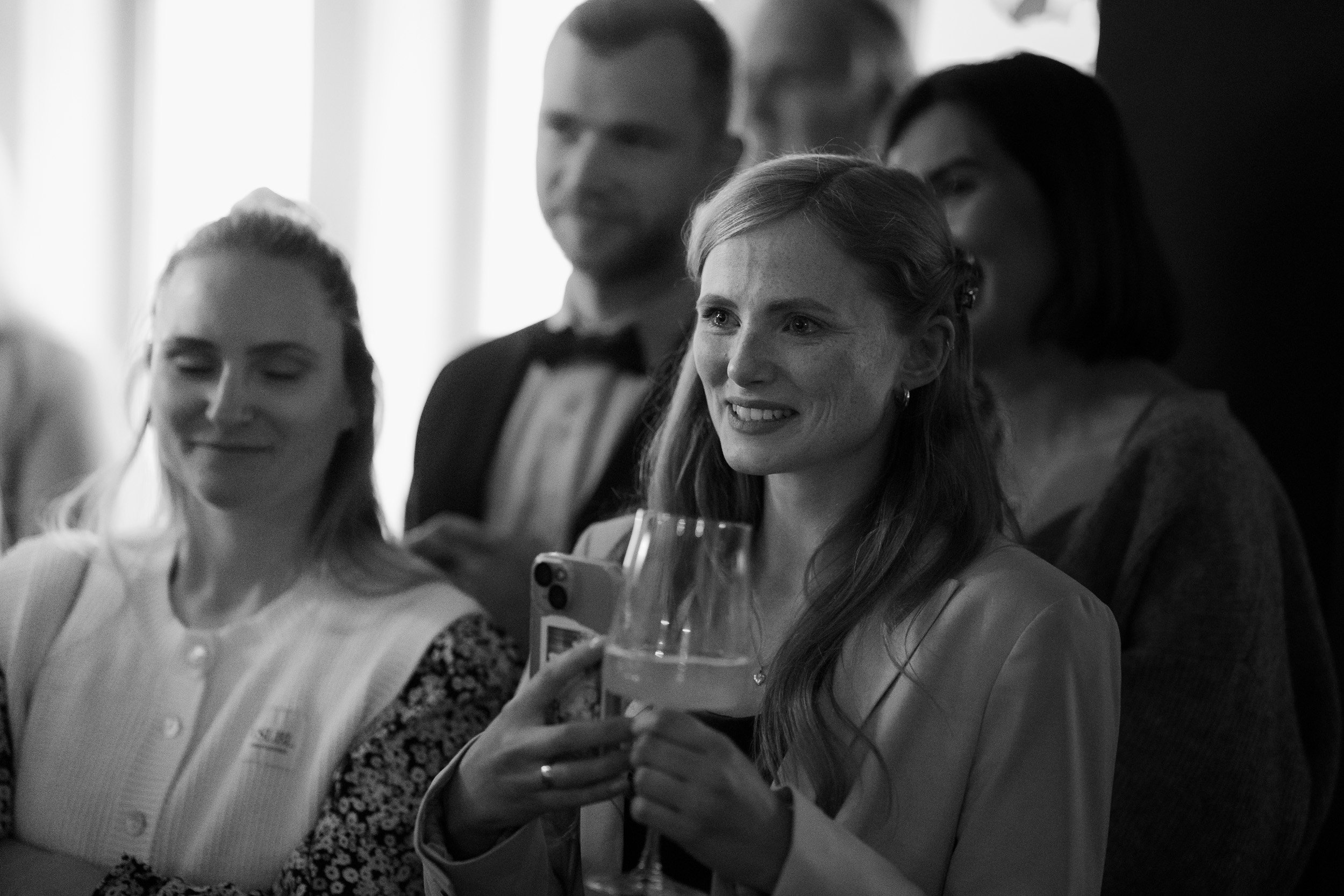 Frau mit langem Haar, die ein Glas Champagner hält und lächelnd bei einer Feier schaut, umgeben von anderen Gästen in eleganter Kleidung.