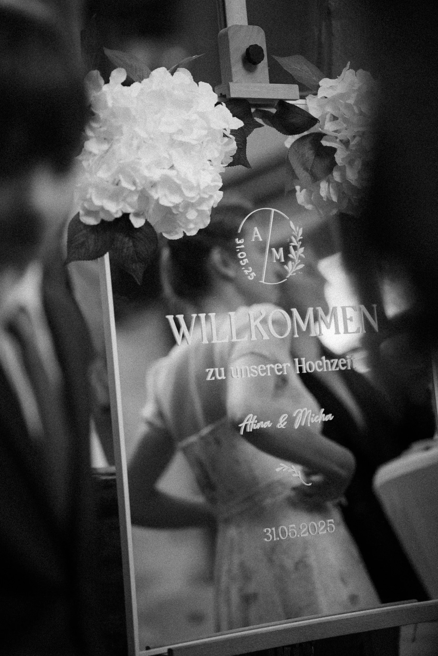 Schwarze Tafel mit deutschem Willkommensgruß zur Hochzeit, Datum 31.05.2025, Namen Anna & Micha, umgeben von weißen Hortensien und grünen Blättern, mit einem kleinen dekorativen Holzobjekt oben.