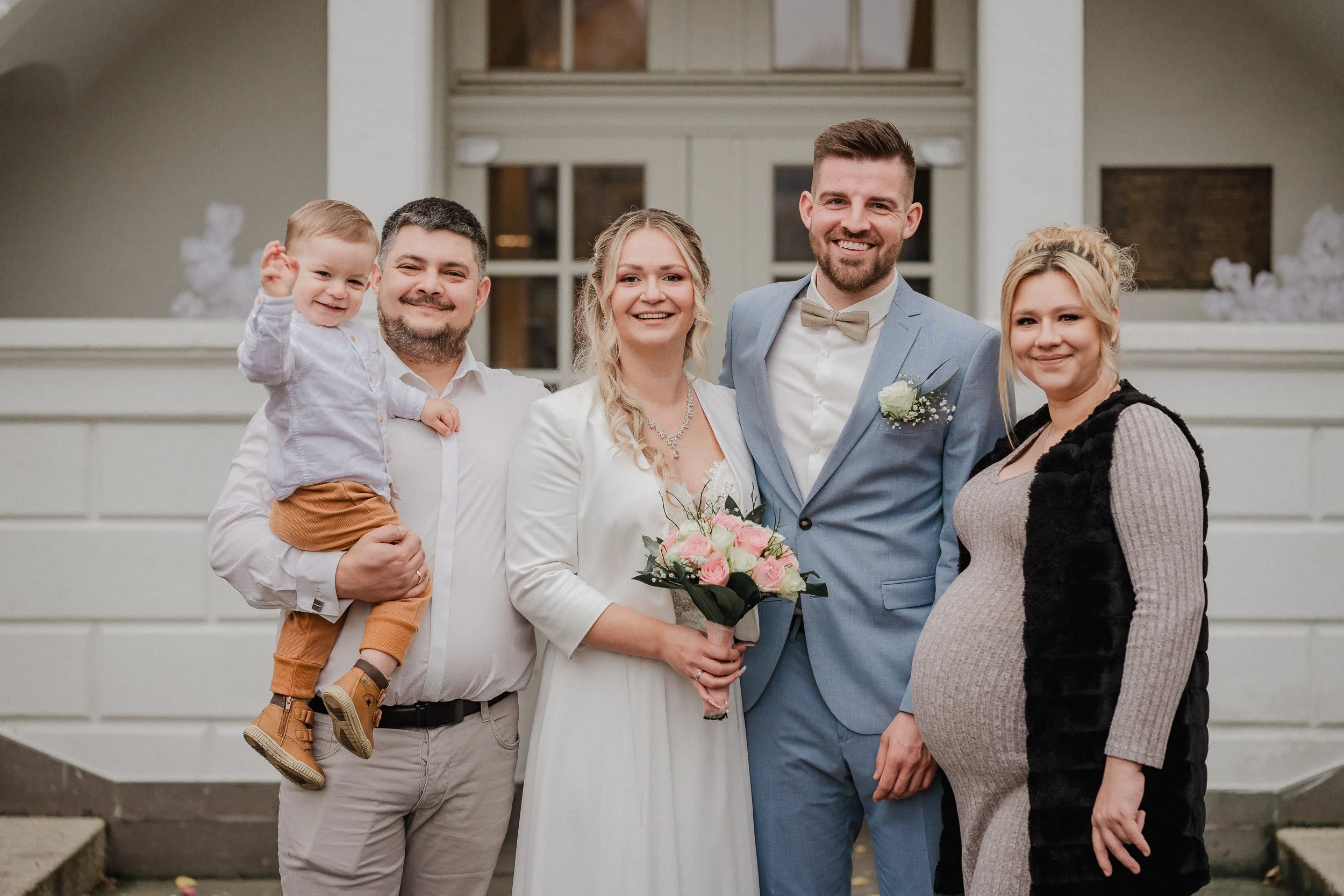 Gruppe von sechs Menschen, darunter ein Baby, bei einer Hochzeitsfeier, mit strahlenden Gesichtern vor einem weißen Gebäude im Freien.