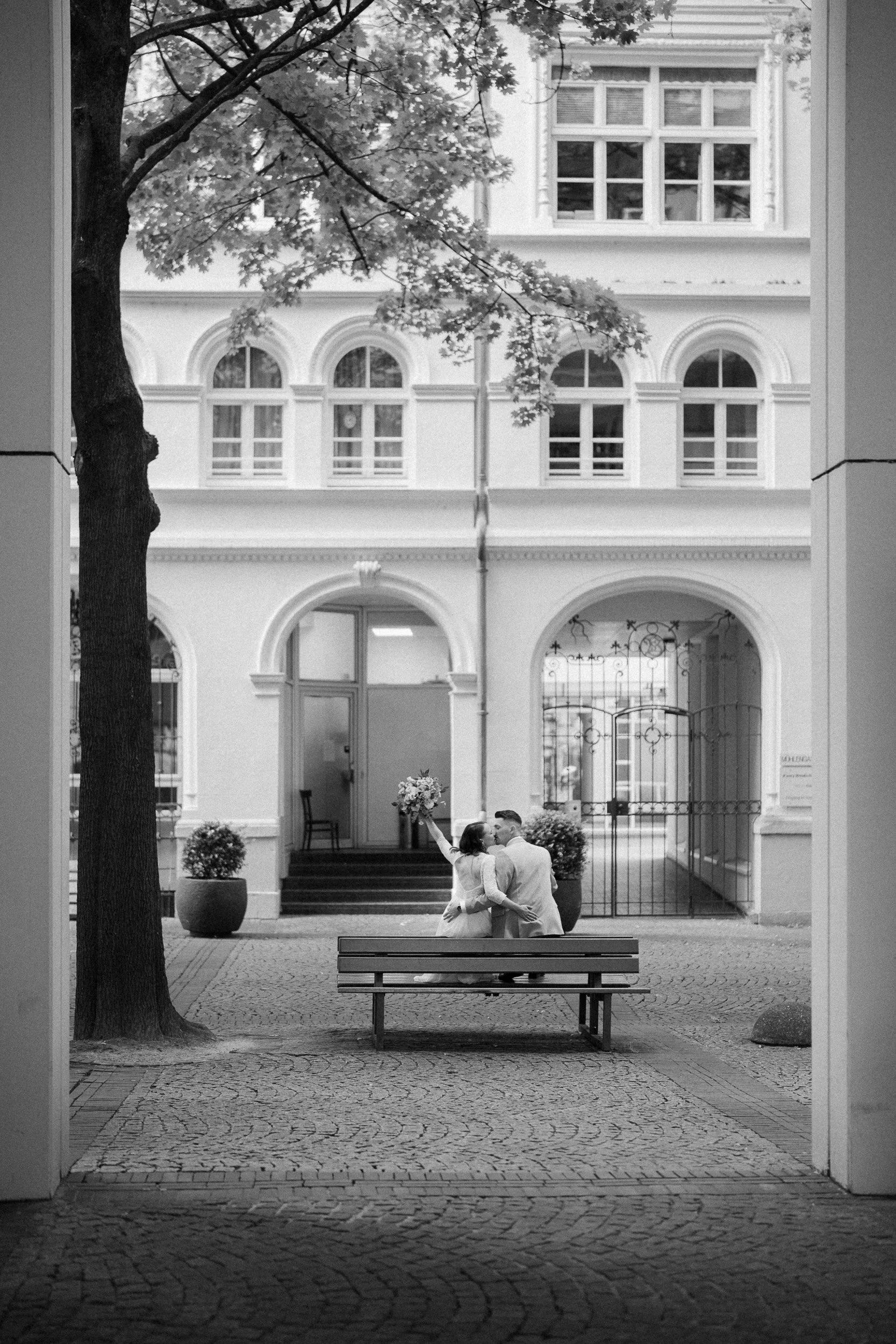 Ein verliebtes Paar, das vor einem historischen Gebäude steht, Kuss küsst und eine Braut, die einen Blumenstrauß hält, im Schwarz-Weiß-Foto.