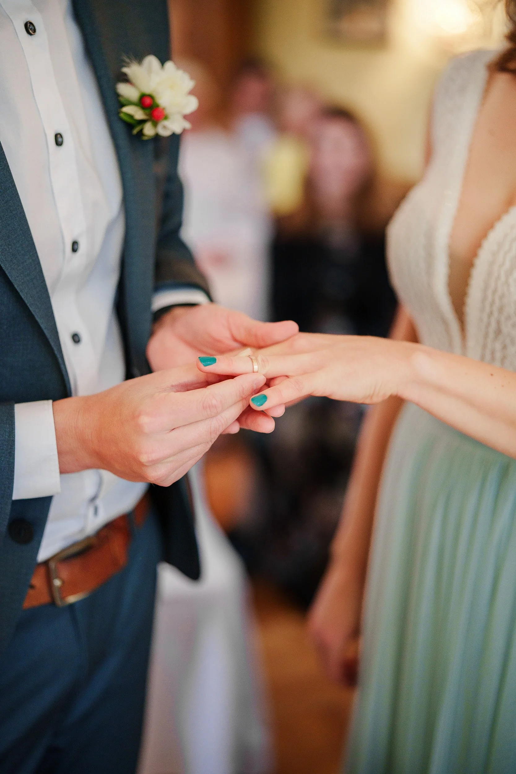 Ein Paar tauscht Eheringe während einer Hochzeitszeremonie, im Hintergrund sind Menschen in einer festlichen Umgebung sichtbar.