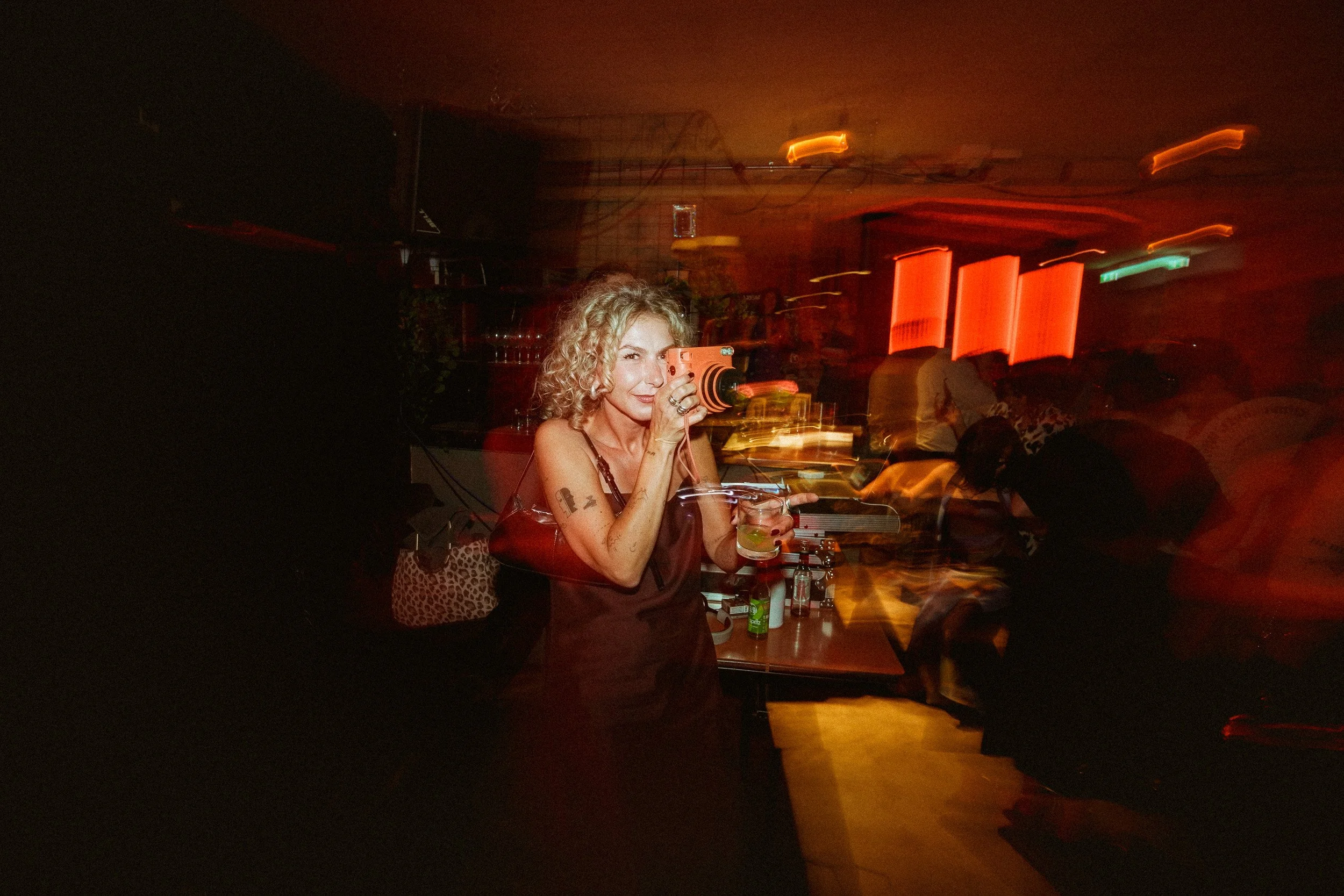 Frau mit lockigem blonde Haar, die in einer Bar mit roten Lichtern photographiert wird, bei Nacht, im dunklen Umfeld, mit Bewegungsunschärfe.