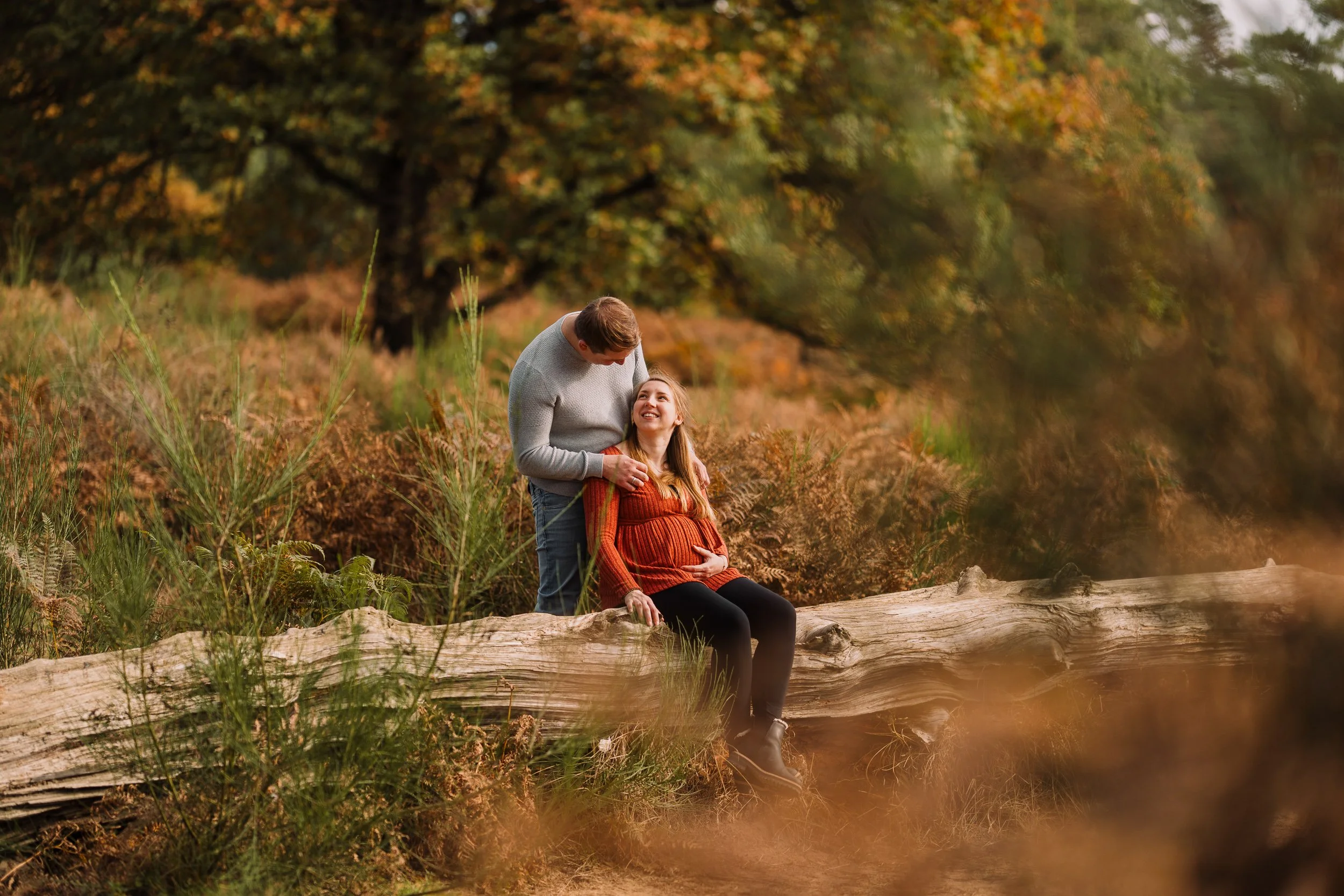 Paar in einer herbstlichen Waldlandschaft, die sich liebevoll um eine schwangere Frau kümmert, die auf einem umgestürzten Baum sitzt.