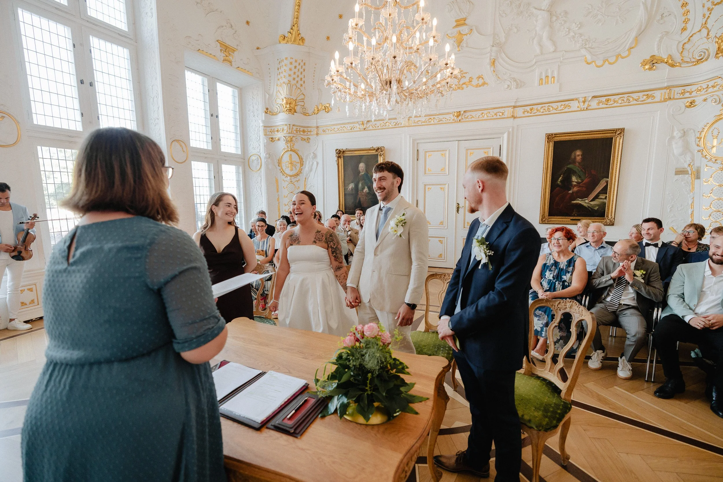 Ein Hochzeitszeremonie im eleganten, aprikfarbenen Raum mit Kronleuchter, bei der das Brautpaar, Trauzeugin und Trauzeuge vor einer Standesbeamtin stehen. Die Gäste sitzen im Hintergrund und nehmen an der Zeremonie teil.
