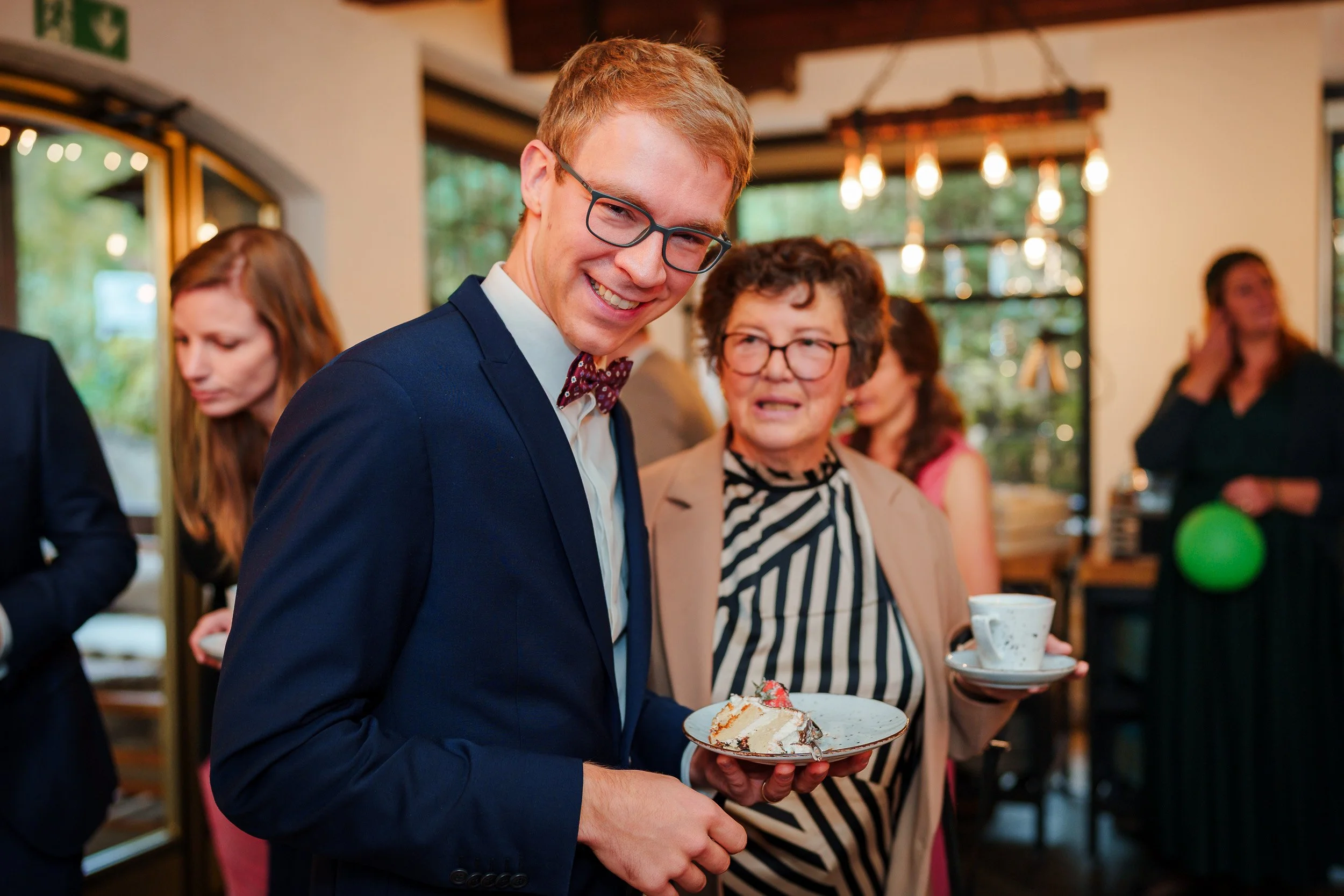 Junger Mann mit Brille, in formeller Kleidung, hält einen Teller mit Kuchen. Ältere Frau neben ihm mit Brille und Striped-Shirt hält eine Tasse. Im Hintergrund weitere Gäste bei einer Feier in einem Raum mit großen Fenstern.