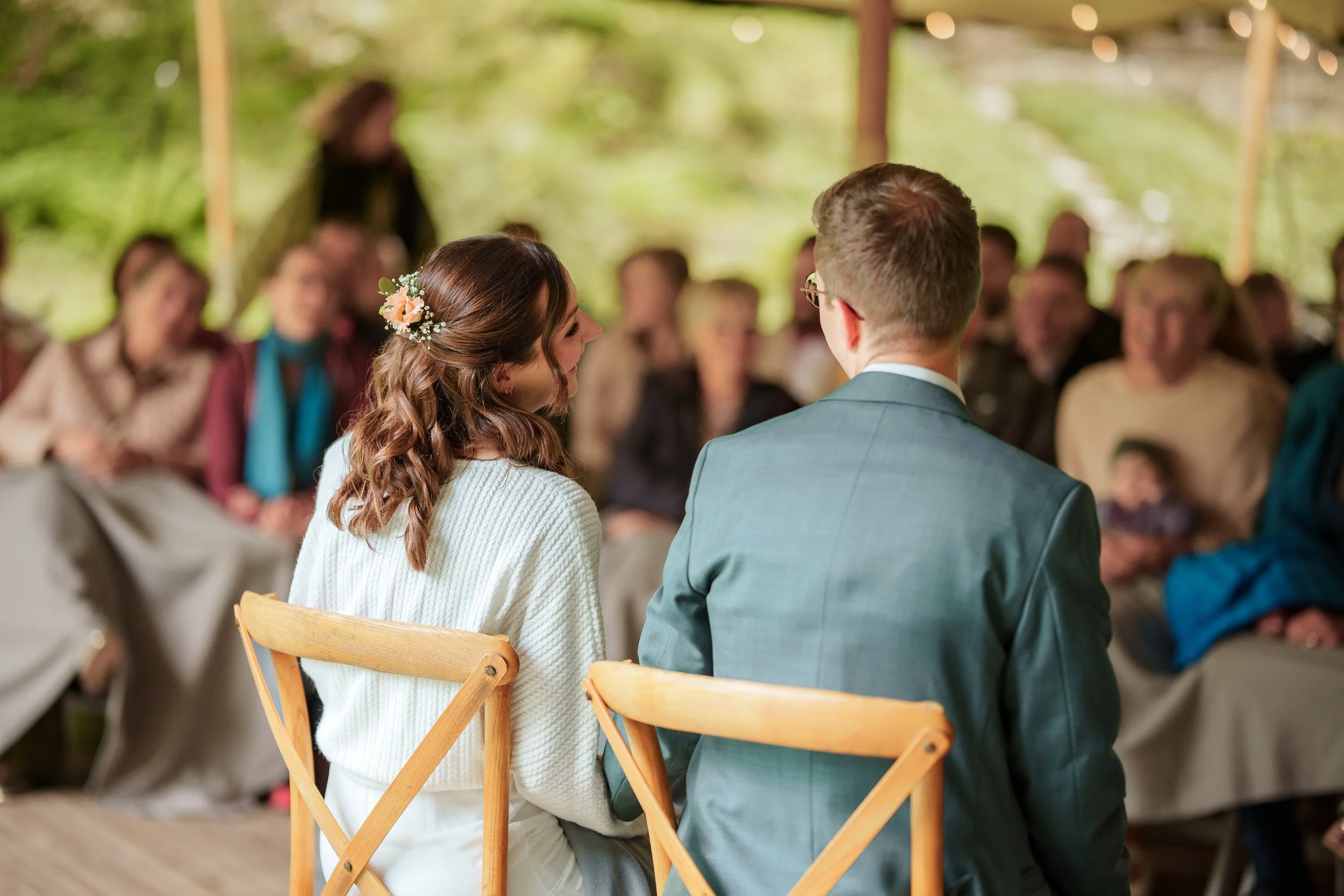 Braut und Bräutigam sitzen bei ihrer Hochzeitszeremonie, umgeben von Gästen in einer outdoor Location mit viel Grün und natürlicher Beleuchtung.