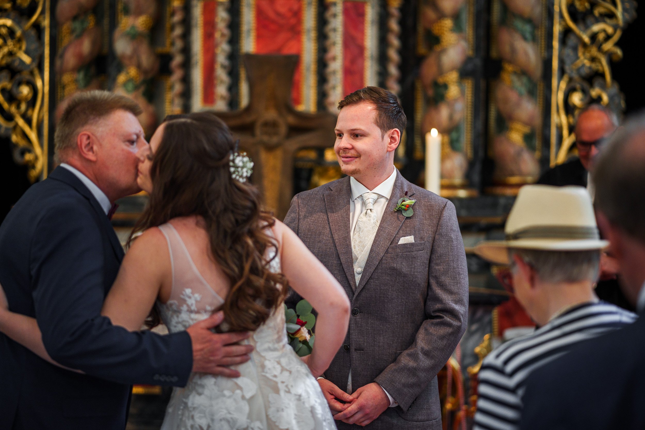 Ein Brautpaar bei einer Zeremonie küsst sich, während der Bräutigam einem anderen Mann in einem schwarzen Anzug zuhört, in einer Kirche mit goldverzierten Details.