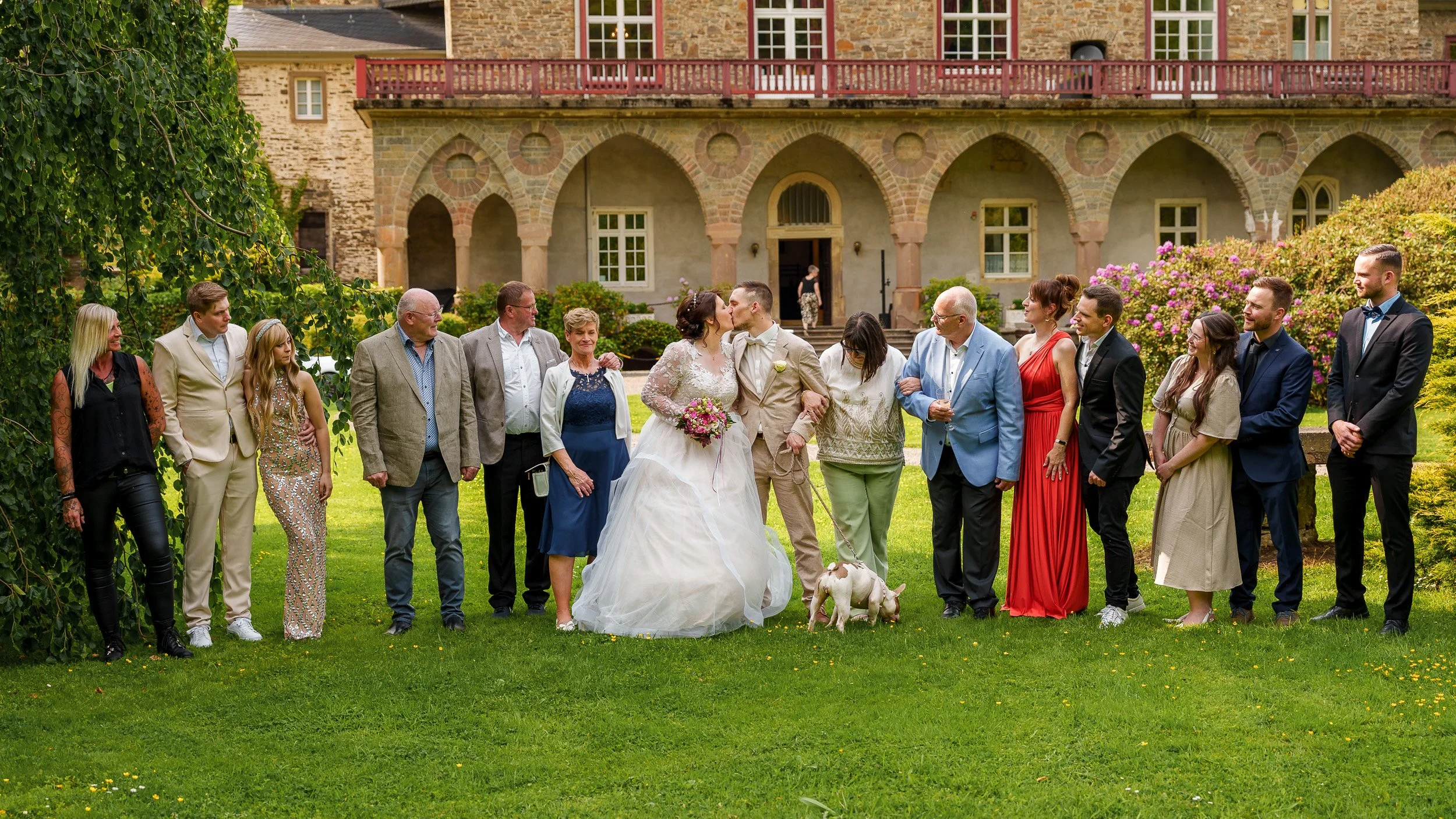 Hochzeitliche Feier im Garten vor einem historischen Gebäude, mit einer Gruppe von Menschen um das Brautpaar, das sich küsst, darunter Familie und Freunde, bei sonnigem Wetter.