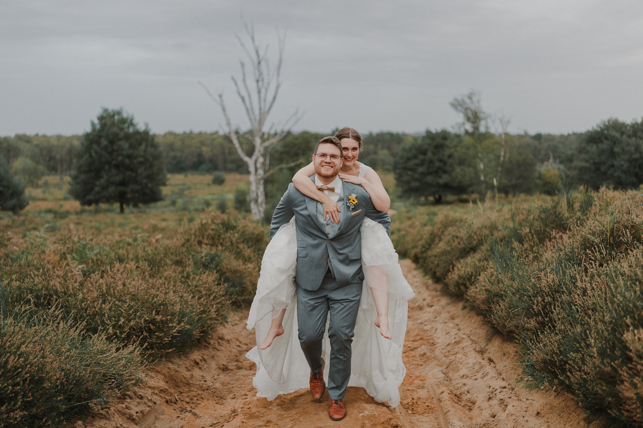 Ein fröhliches Paar im Hochzeitsoutfit, der Bräutigam trägt einen grauen Anzug und der Brautkleid, der Bräutigam trägt Brille, trägt eine Sonnenblume im Revers, der Himmel ist bewölkt, sie laufen auf einer sandigen Landstraße durch eine ländliche Lan