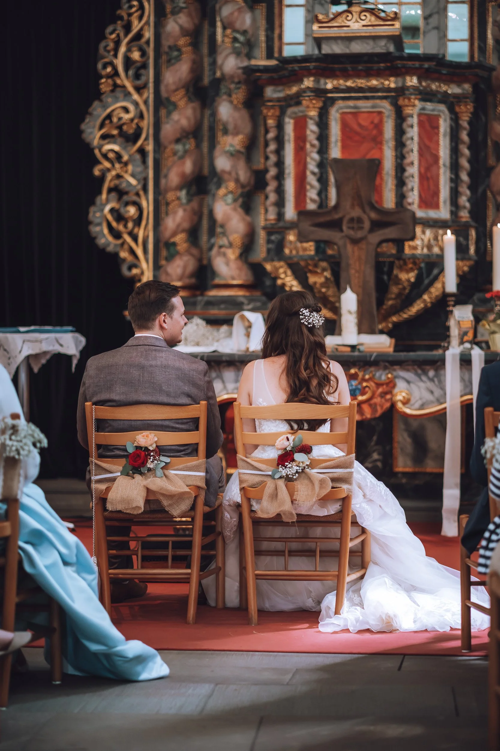 Brautpaar bei einer Zeremonie in einer Kirche, sitzend auf Stühlen mit Blumen und Bändern. Im Hintergrund ein Altar mit Kerzen, Kreuz und kunstvollen Verzierung.