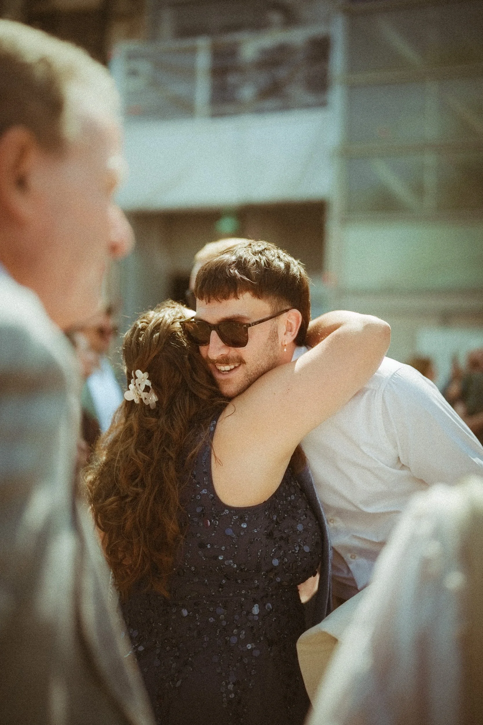 Ein Mann mit dunklen Haaren, Sonnenbrille und Ohrring umarmt eine Frau bei einem sonnigen Treffen, im Hintergrund sind andere Menschen.