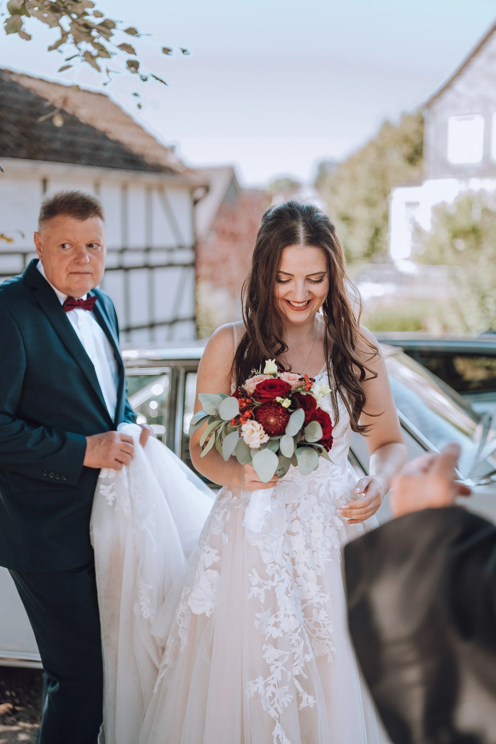 Junge Frau in Brautkleid mit Brautstrauß, älterer Mann in Anzug und Krawatte, vermutlich Vater, bei einer Hochzeit, draußen neben einem Auto