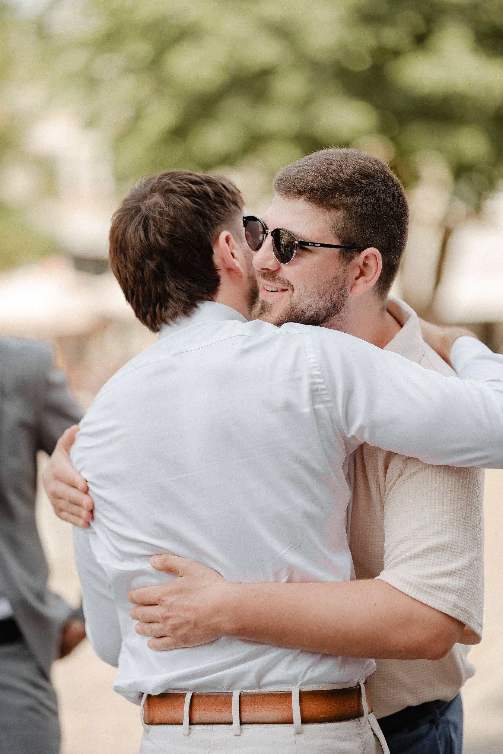 Zwei Männer umarmen sich, einer trägt Sonnenbrille, im Hintergrund sind Bäume und eine weitere Person in formeller Kleidung.
