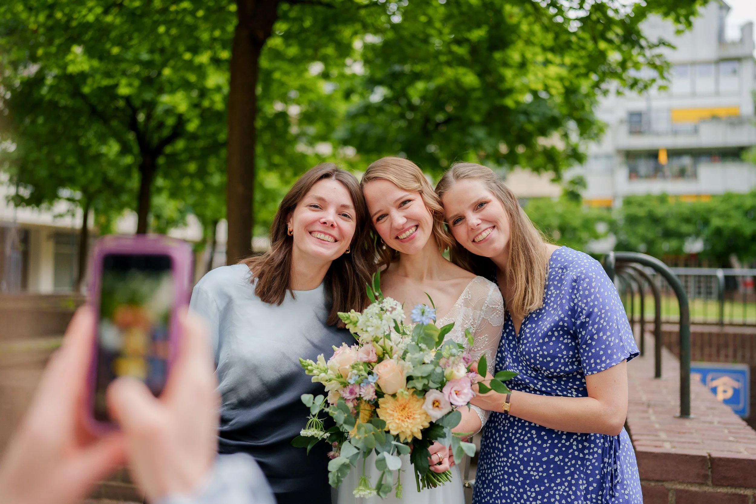 Drei lachende Frauen beim Fotografieren, eine hält einen Blumenstrauß, im Park bei grünen Bäumen und Gebäuden im Hintergrund.