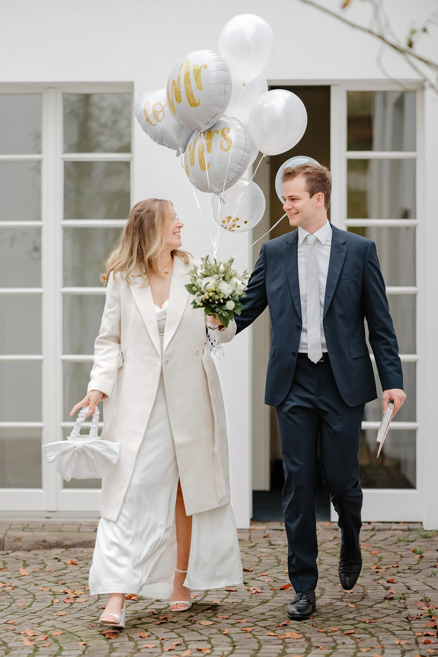 Ein glückliches Brautpaar läuft gemeinsam vor einem weißen Haus mit großen Fenstern. Die Braut trägt ein weißes Kleid und einen cremefarbenen Mantel, hält einen weißen Blumenstrauß und Ballons, auf denen 'Mrs' und 'Love' geschrieben steht. Der Bräuti