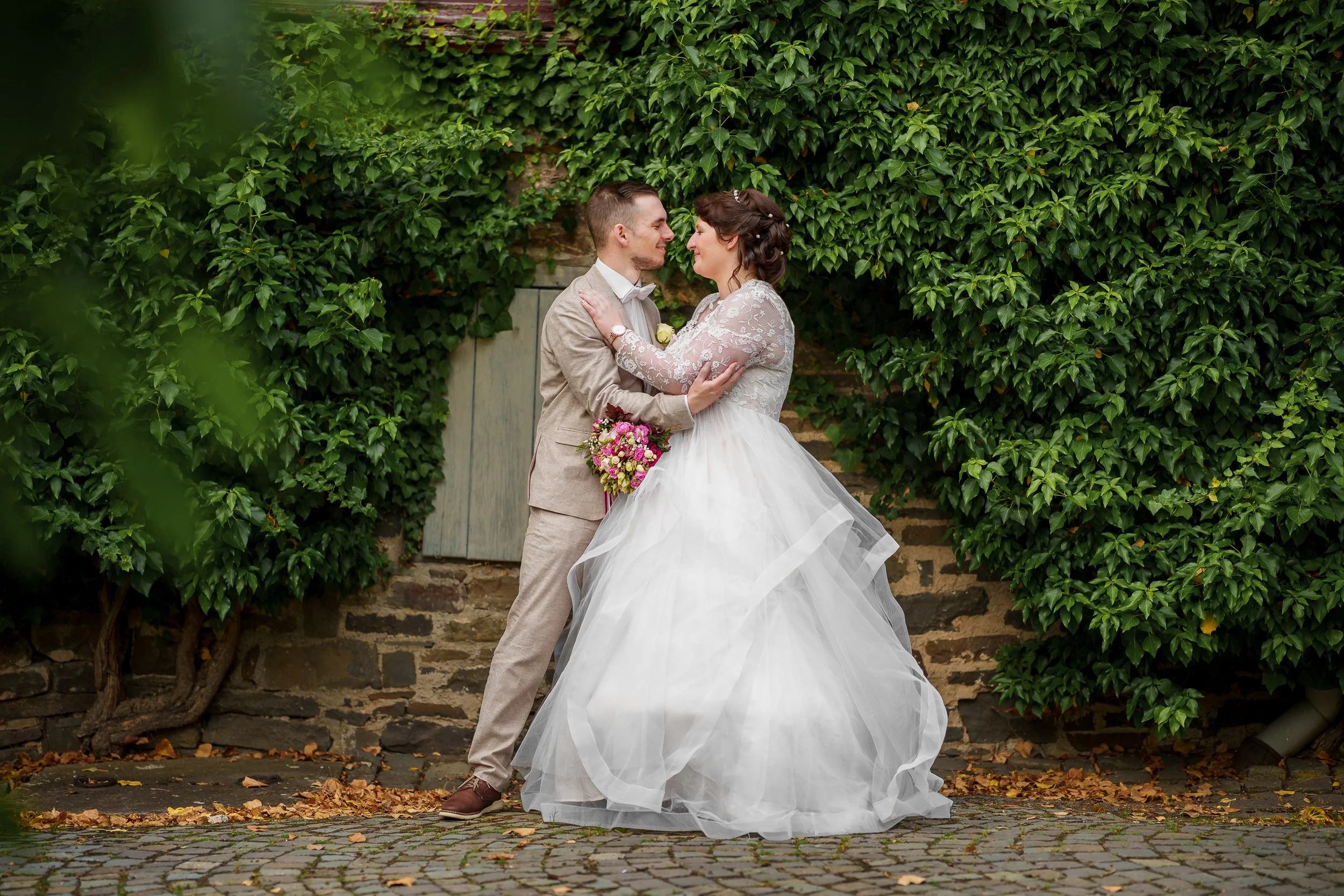 Ein Brautpaar steht sich gegenüber vor einer mit grünen Efeu bewachsenen Mauer, der Bräutigam trägt einen hellen Anzug und die Braut ein weißes Hochzeitskleid, sie hält einen Blumenstrauß in der Hand, beide schauen sich verliebt an.