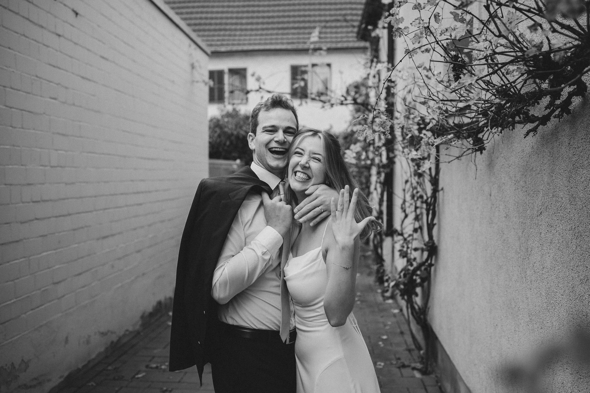 Ein glückliches Paar in festlicher Kleidung, das sich umarmt und lacht, in einer schmalen Gasse mit einer bemalten Mauer und Blumen auf der rechten Seite, Schwarz-Weiß-Fotografie.