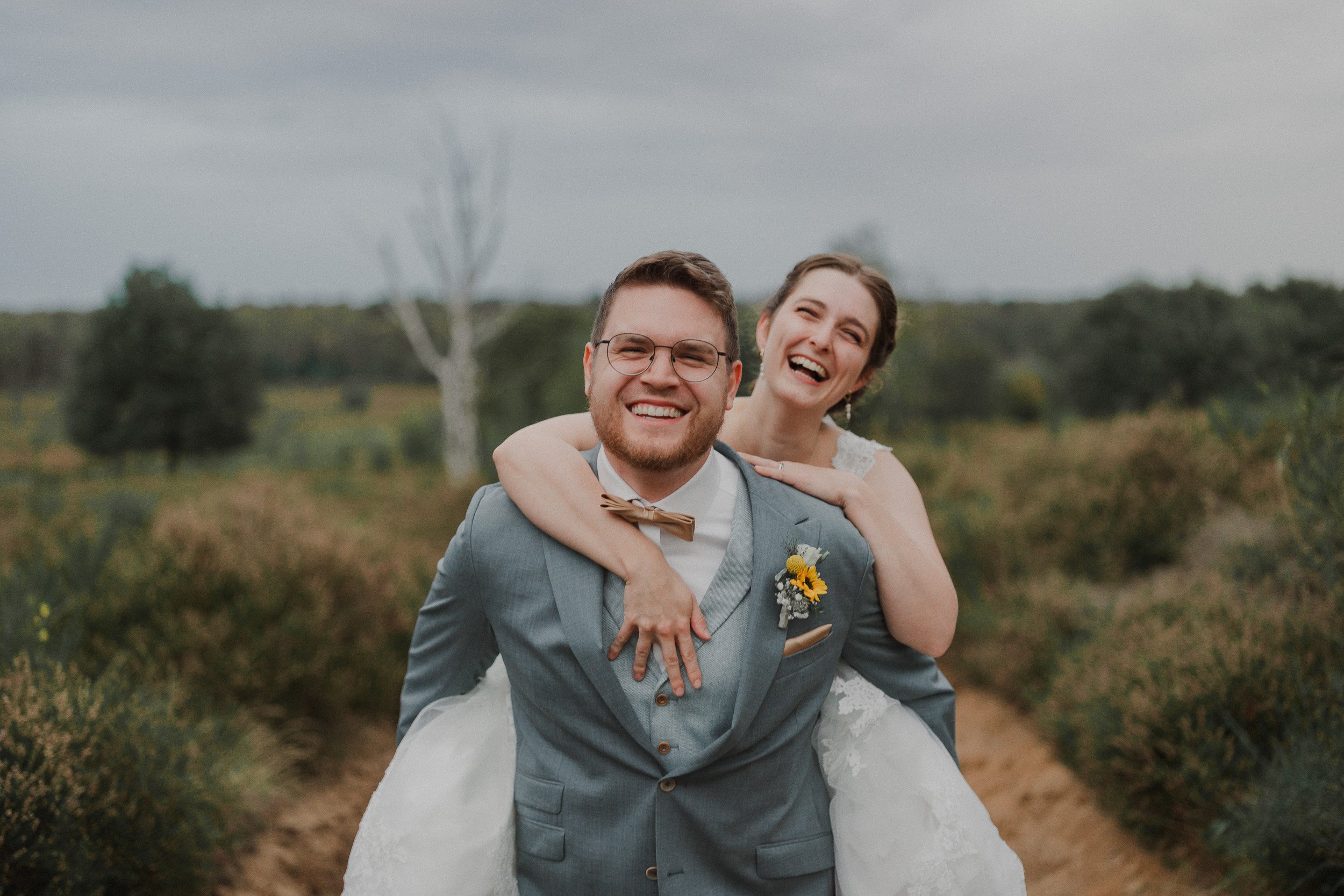Ein glückliches Paar bei einer Hochzeit, der Mann trägt einen grauen Anzug mit einer Sonnenblumen-Brosche, die Frau trägt ein weißes Hochzeitskleid und sitzt auf seinem Rücken in einer natürlichen, grünen Landschaft mit Bäumen.
