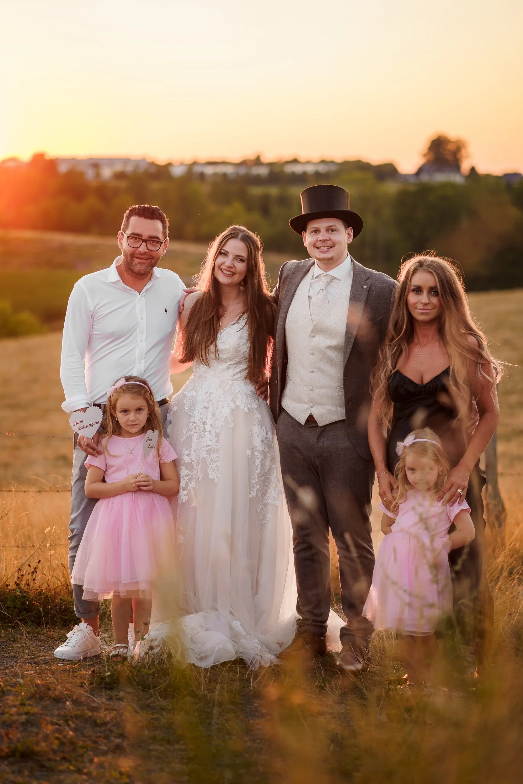 Eine Gruppe von sechs Menschen, darunter Mädchen in pinken Kleidern, posiert beim Sonnenuntergang in einer offenen Landschaft.