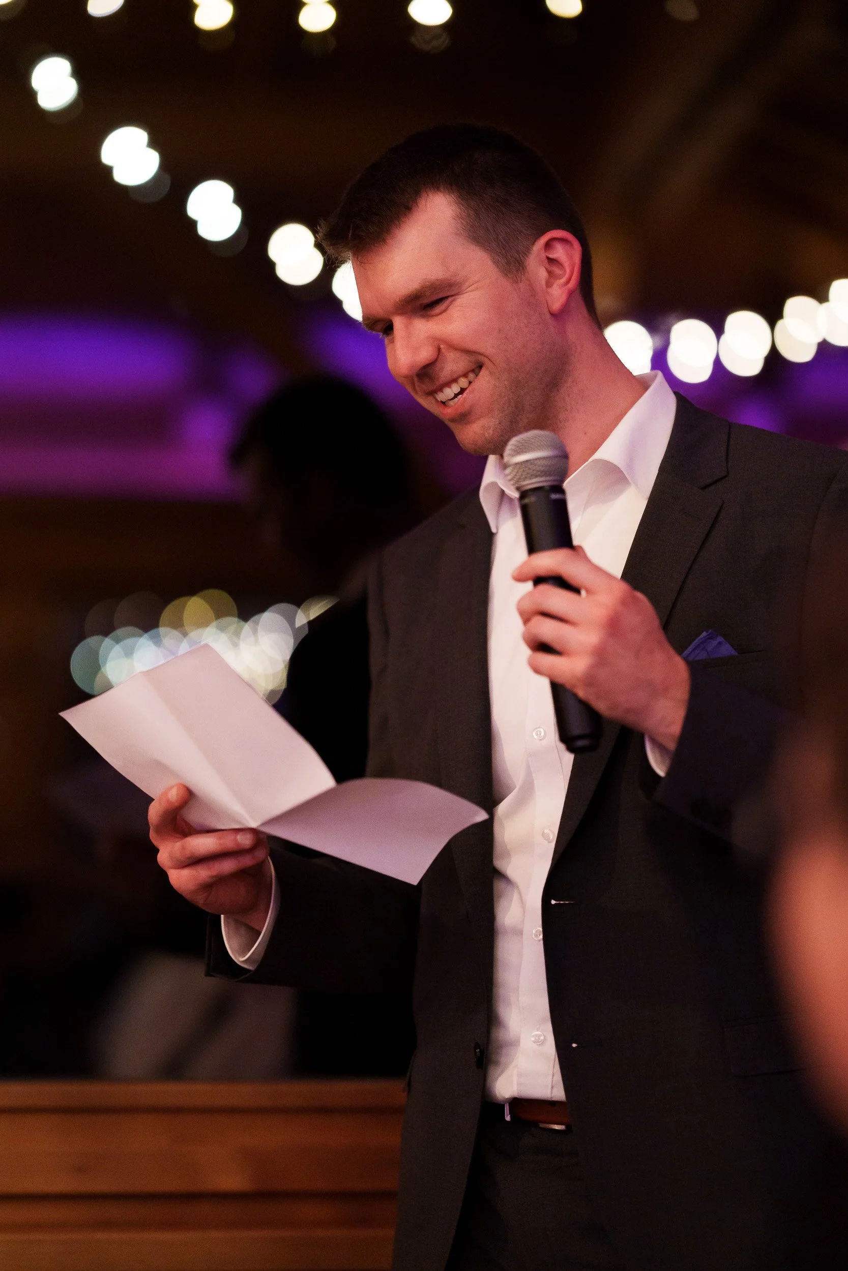 Ein junger Mann in einem dunklen Anzug liest bei einer Veranstaltung aus einem Papier, während er mit einem Mikrofon spricht. Der Hintergrund ist mit bunten Lichterdekorationen beleuchtet.