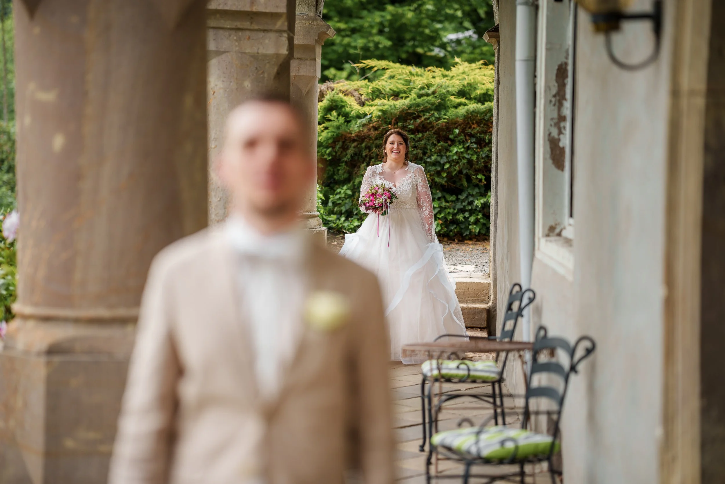 Braut im weißen Kleid hält einen Blumenstrauß, im Hintergrund steht der Bräutigam, beide bei einer Hochzeitsaufnahme im Freien.