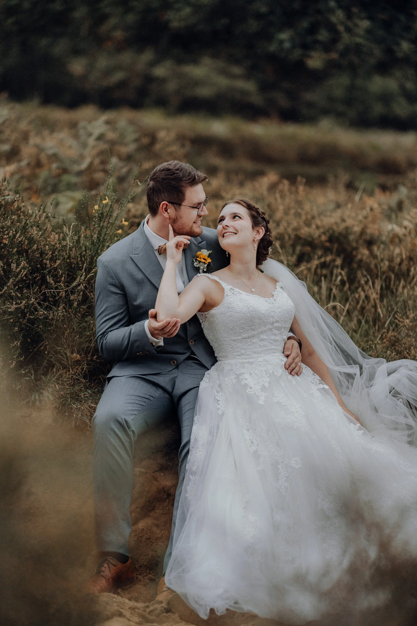 Ein Brautpaar sitzt im Freien, der Bräutigam in einem grauen Anzug und die Braut in einem weißen Hochzeitskleid, schauen sich liebevoll an.