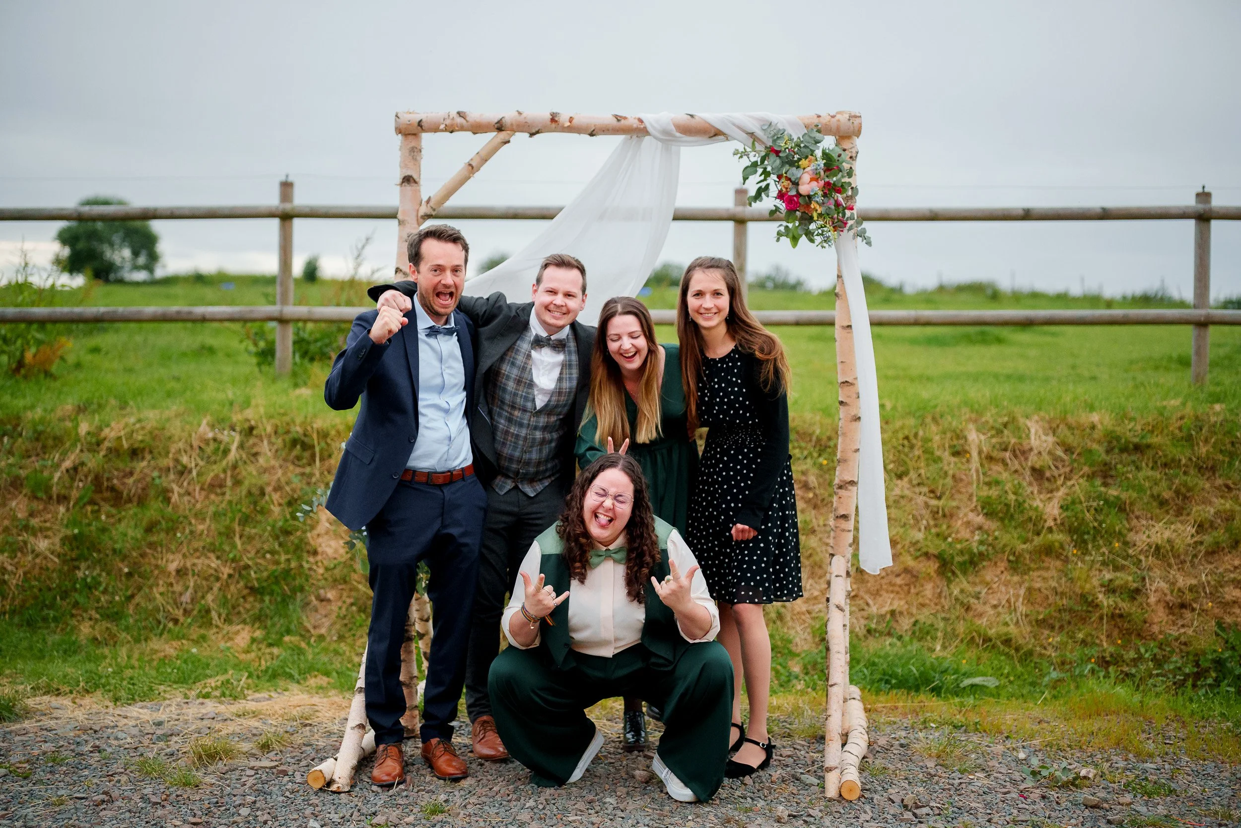 Menschen feiern bei einer Hochzeit im Freien vor einem Holzbogen mit Blumen. Sie lachen und posieren für das Foto.