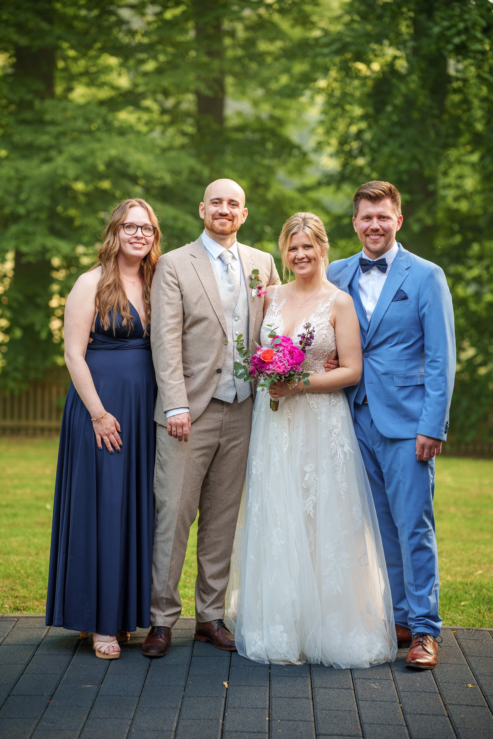 Gruppenfoto bei einer Hochzeit mit geschmücktem Brautpaar und zwei Gästen vor einem grünen Baumhintergrund.