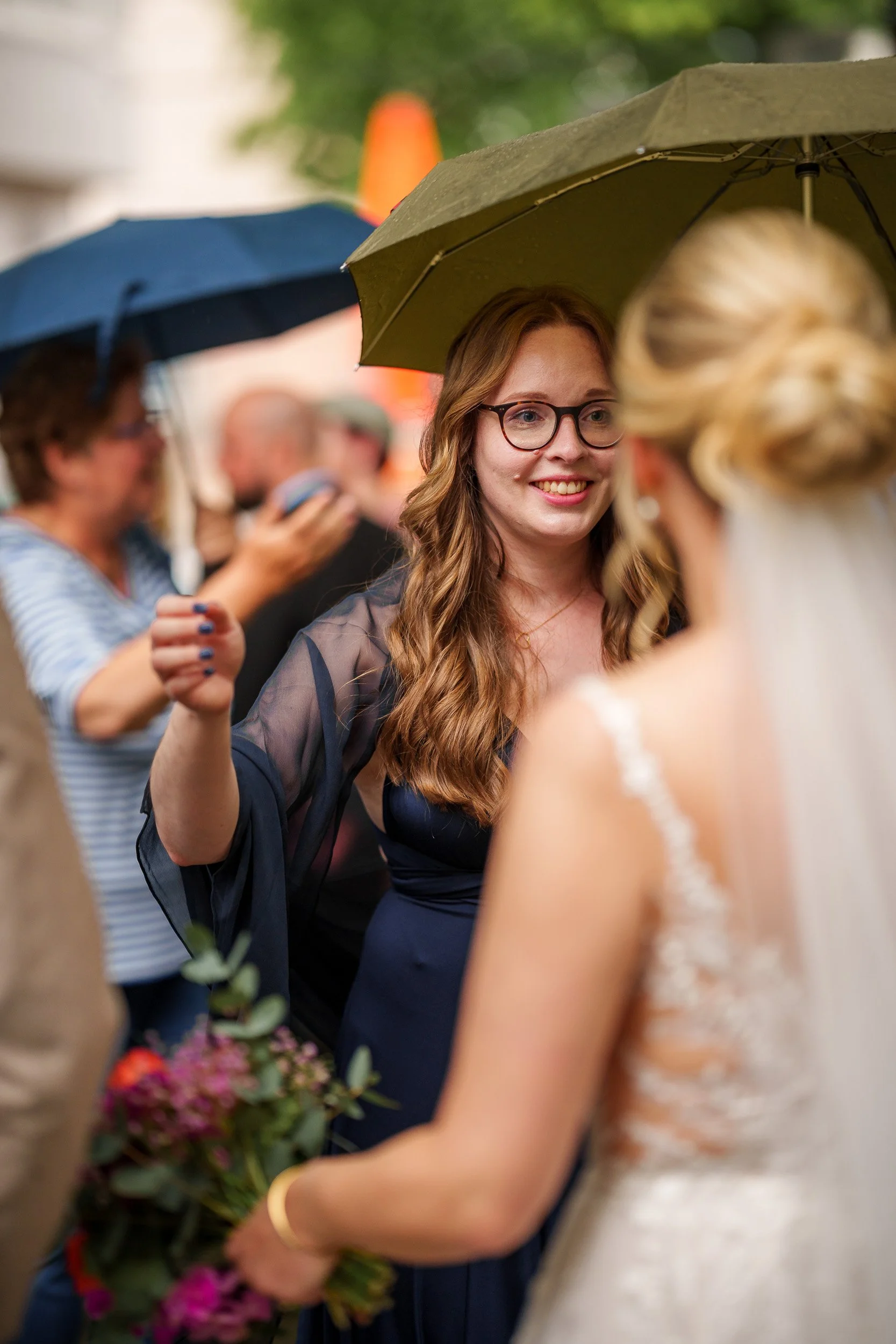 Frau mit Brille und langen, lockigen Haaren lächelt bei einer Hochzeit im Regen, während sie einer Braut mit Blumen in der Hand zuhört.