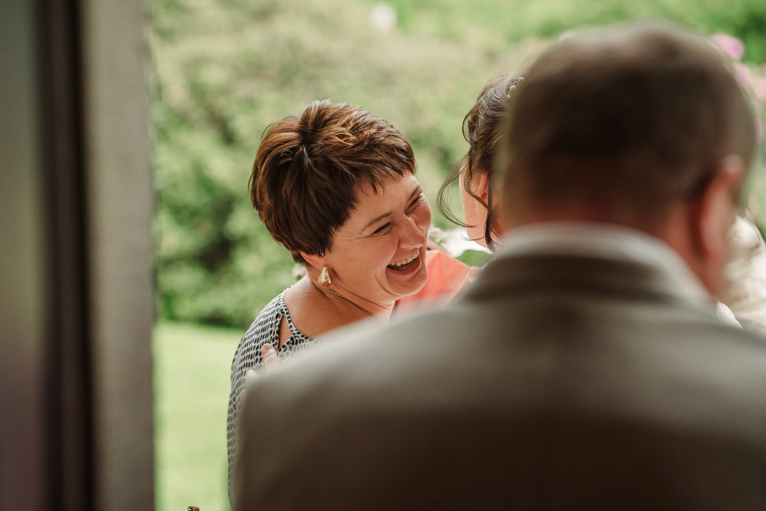 Eine lachende Frau mit kurz braunen Haaren und goldenen Ohrringen, die mit anderen Menschen bei einem Treffen im Freien sitzt.