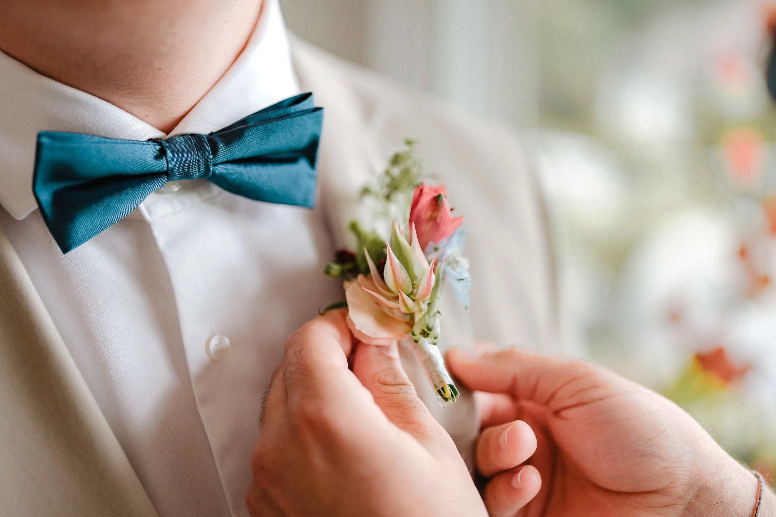 Nahaufnahme einer Person, die ein Blumenanstecker am Revers eines weißen Anzugs anbringt, mit einer blauen Schleife am Hals, vermutlich bei einer Hochzeitsfeier.