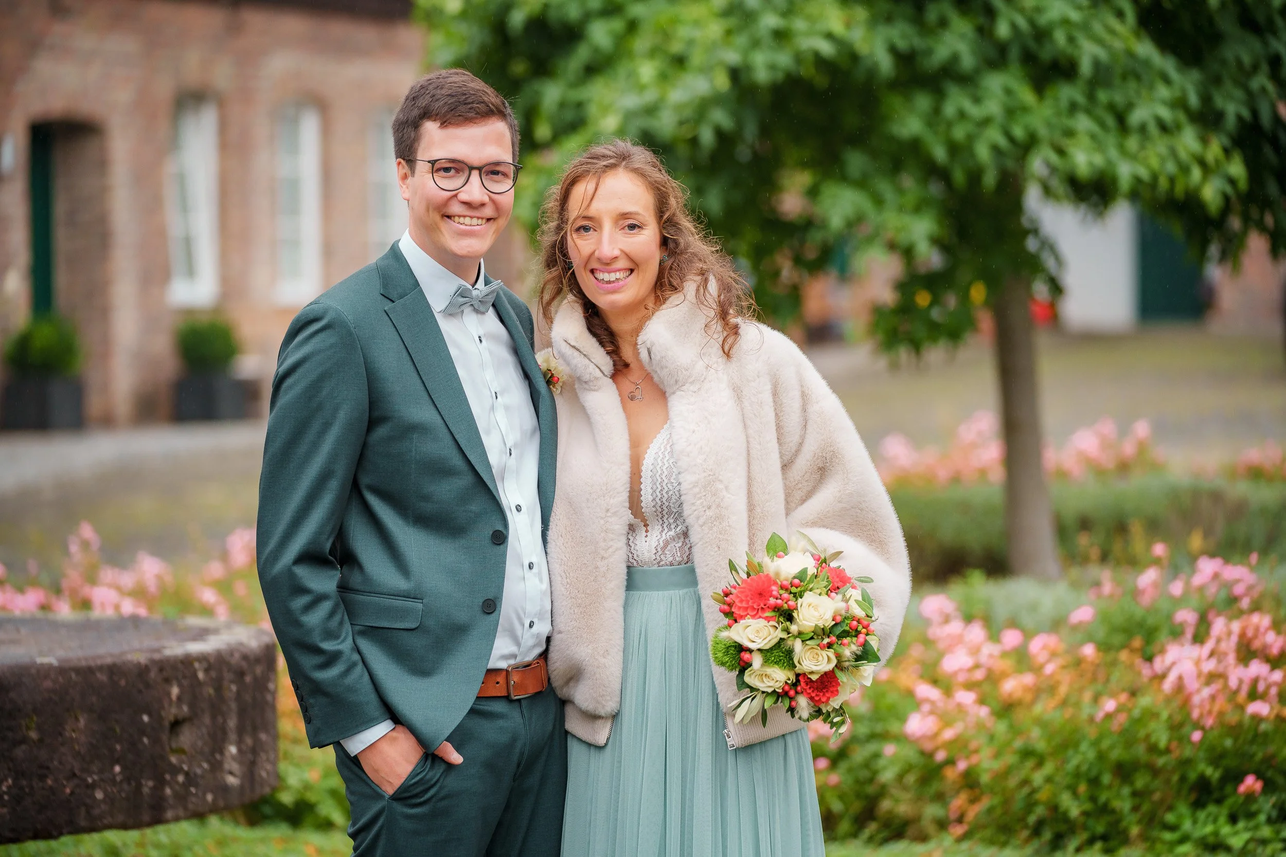 Ein verliebtes Paar steht im Grünen, die Frau hält einen Blumenstrauß, beide lächeln, im Hintergrund Bäume und Blumen. Es sieht aus wie eine Hochzeitsaufnahme.