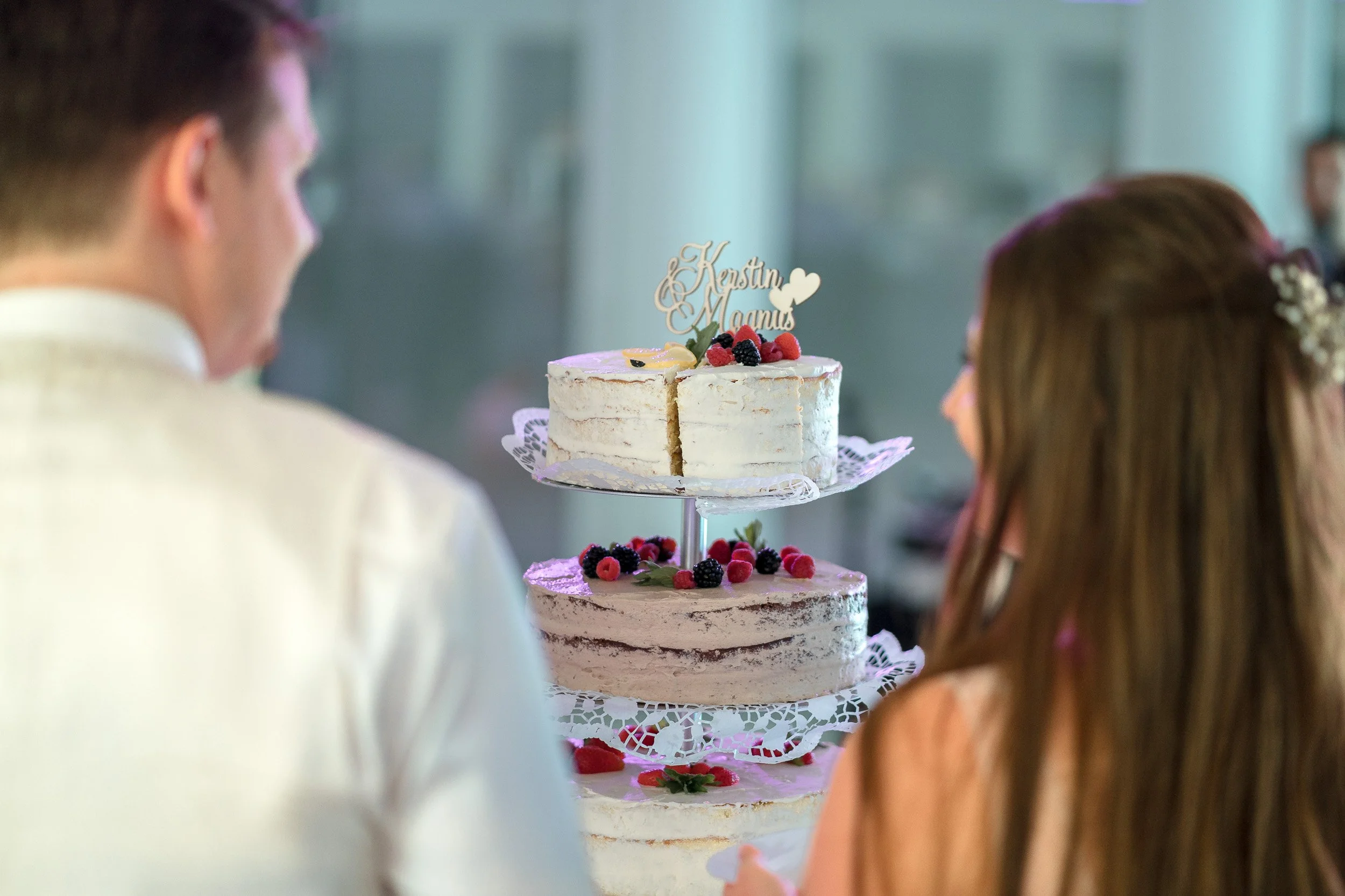Ein Brautpaar blickt auf eine dreistöckige Hochzeitstorte, dekoriert mit Beeren und einem Holz-Hochzeitskuchen-Topper.