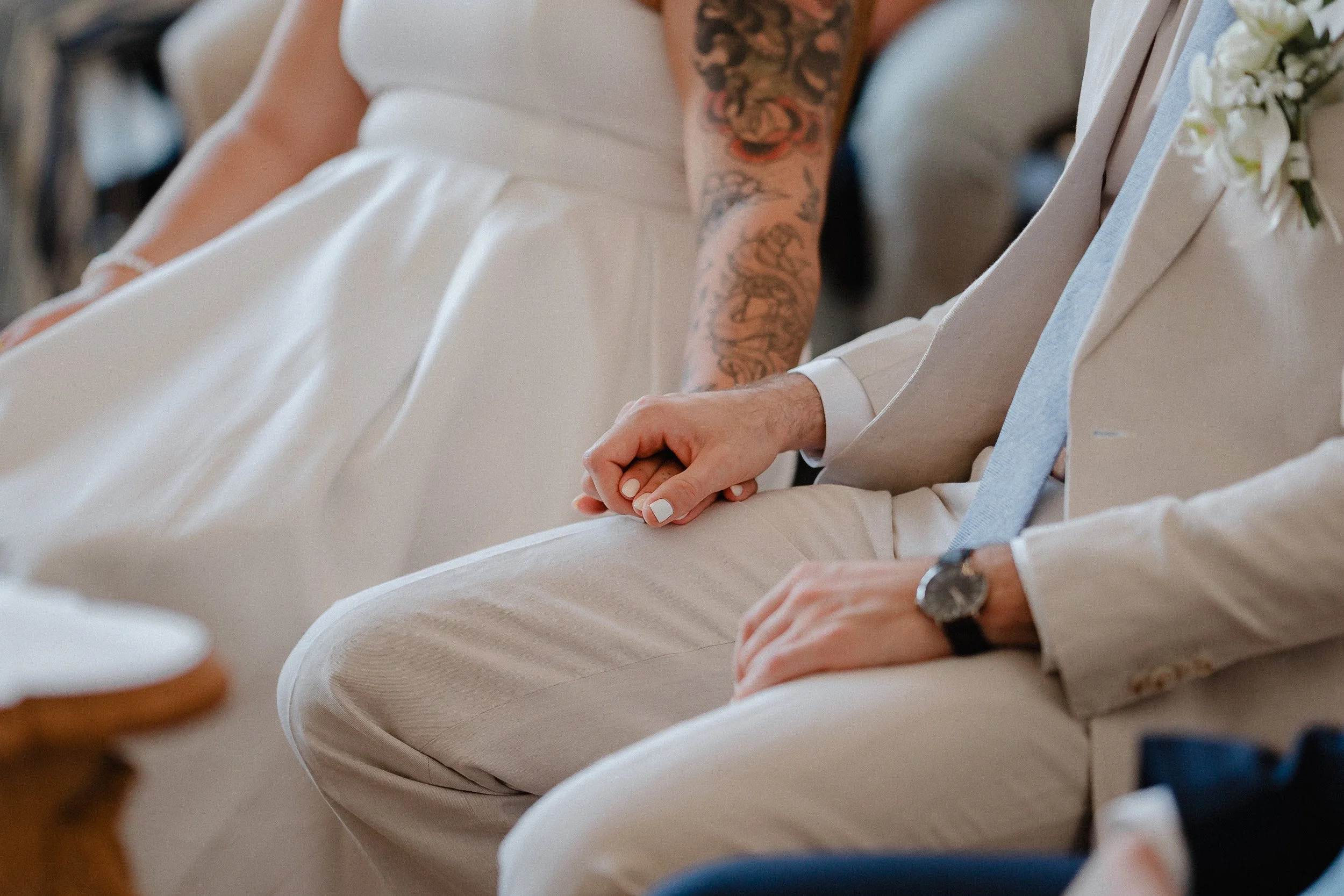 Nahaufnahme von einem Brautpaar, das bei einer Hochzeitszeremonie Hände hält, beide in eleganten weißen Anzügen, der Mann trägt eine Uhr, die Frau hat eine Tätowierung am Arm.
